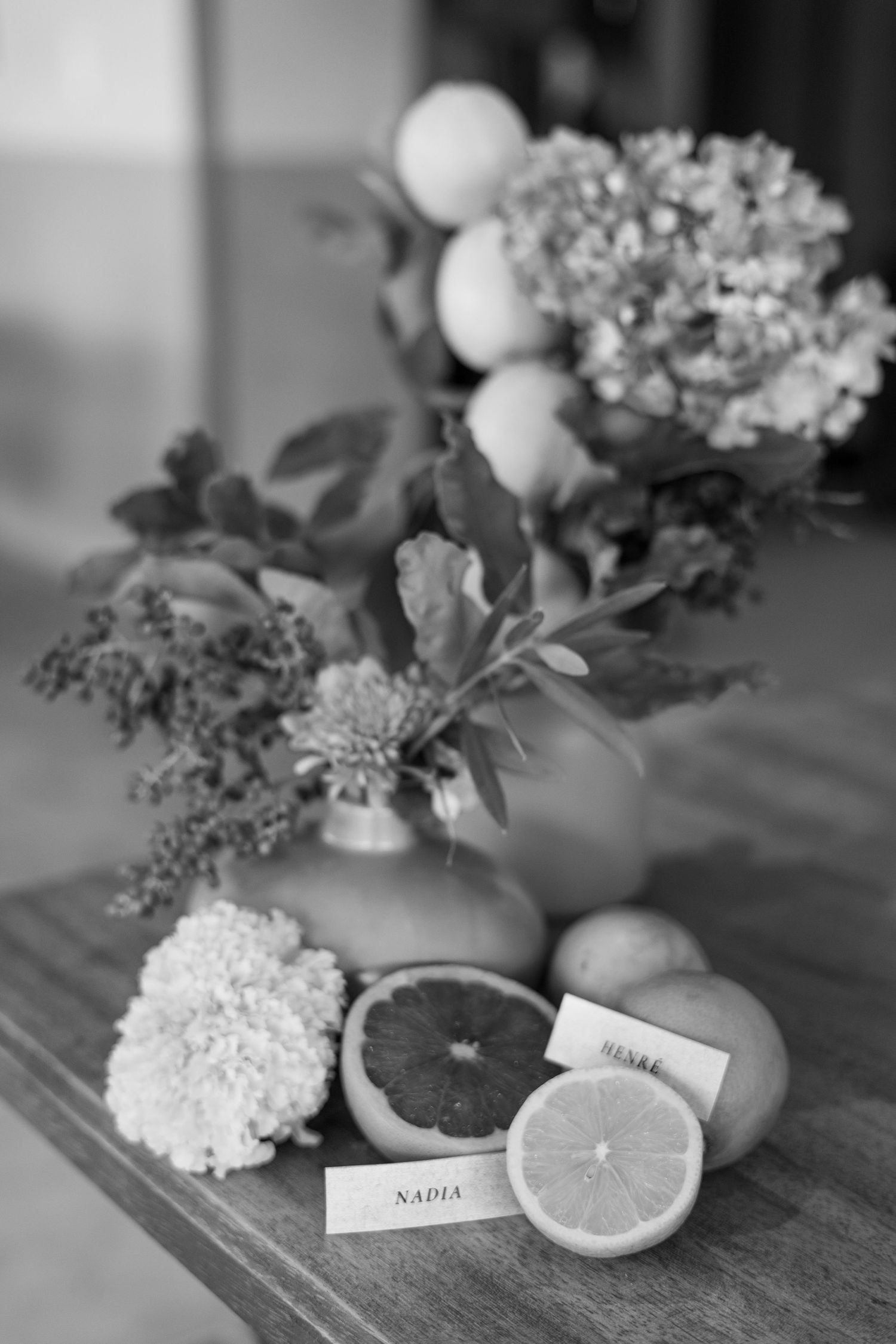 Black and white still life with citrus slices, white flowers, and name cards on a wooden surface.