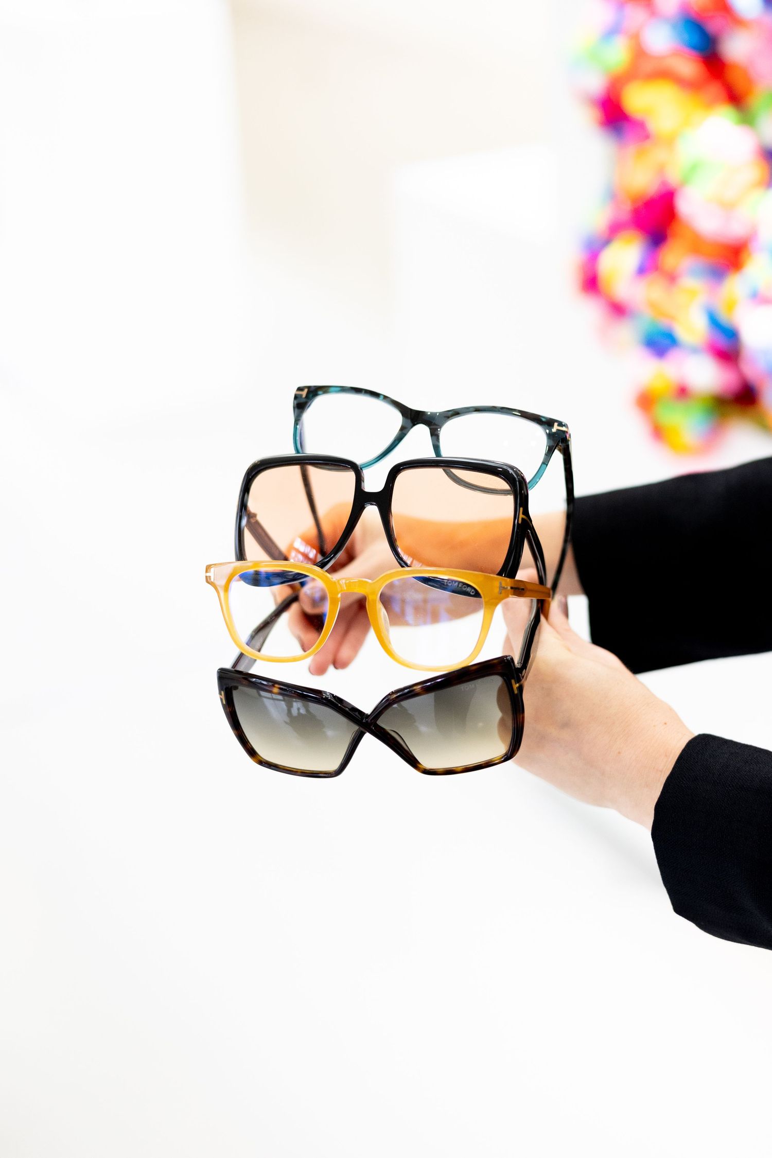 Hands holding three stylish eyeglass frames in different styles against a white background with colorful blur.