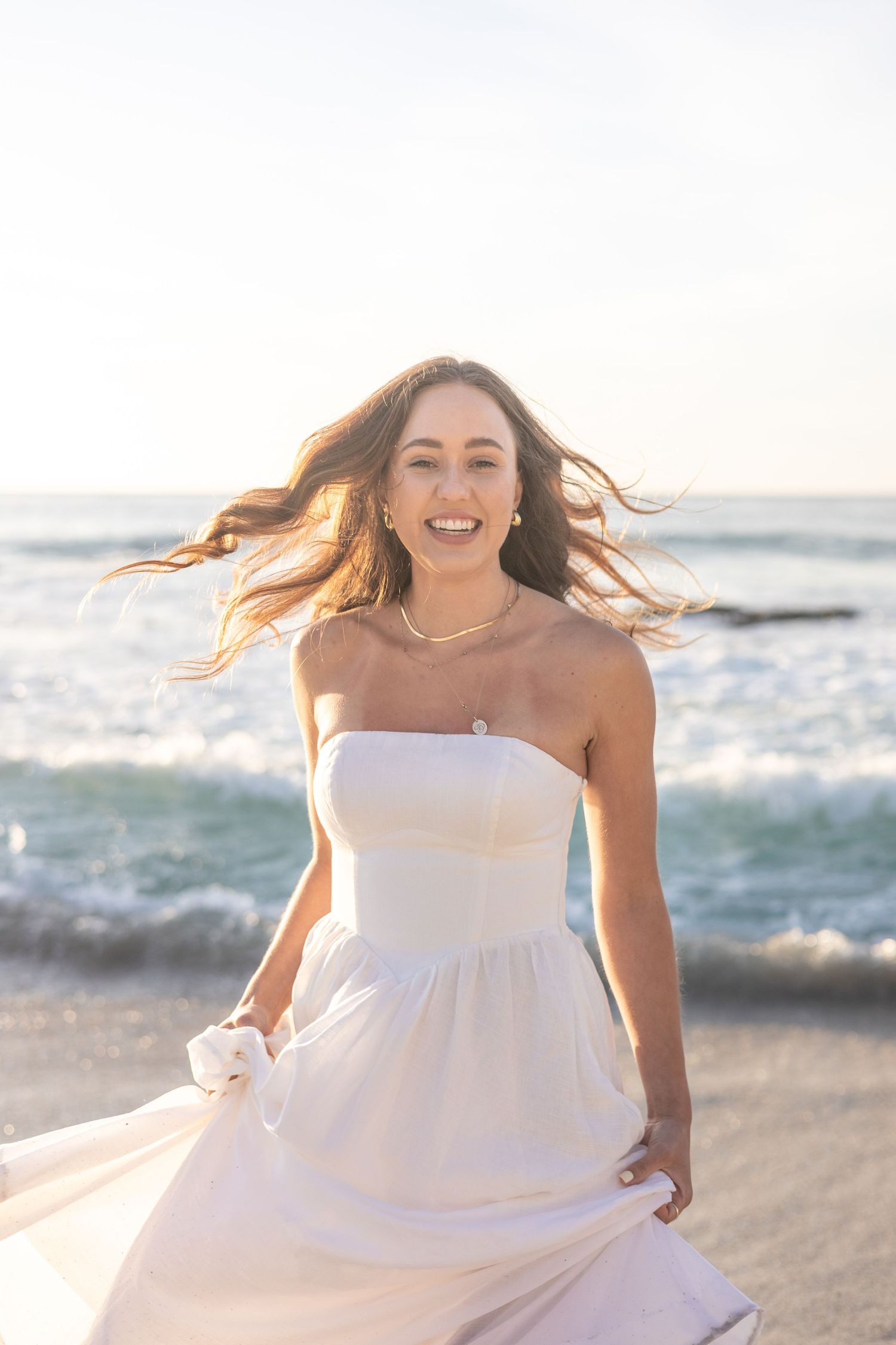 Candid beach bridal portrait with windswept hair and flowing white dress at sunset.
