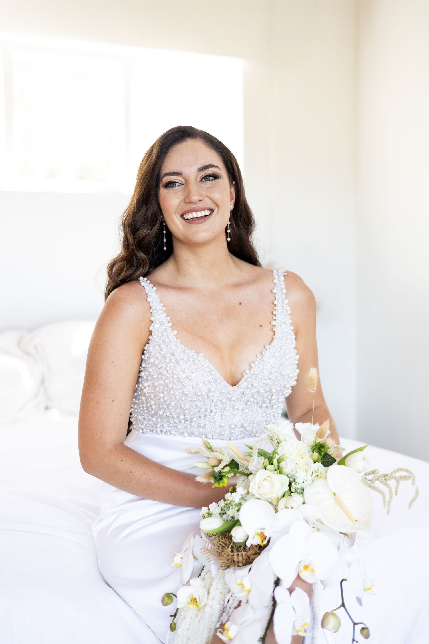 A bride wearing a white beaded V-neck gown sits holding an elegant white and green floral bouquet against a bright backdrop.