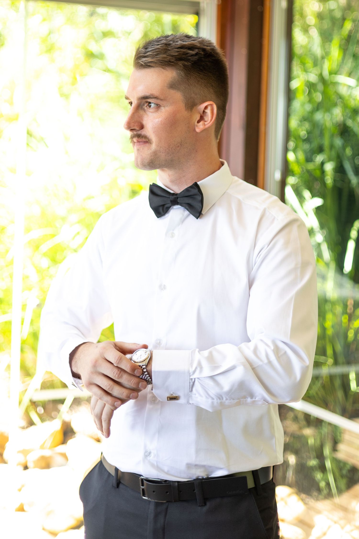 Well-dressed person in formal attire with white dress shirt, black bow tie and dark pants standing by window.