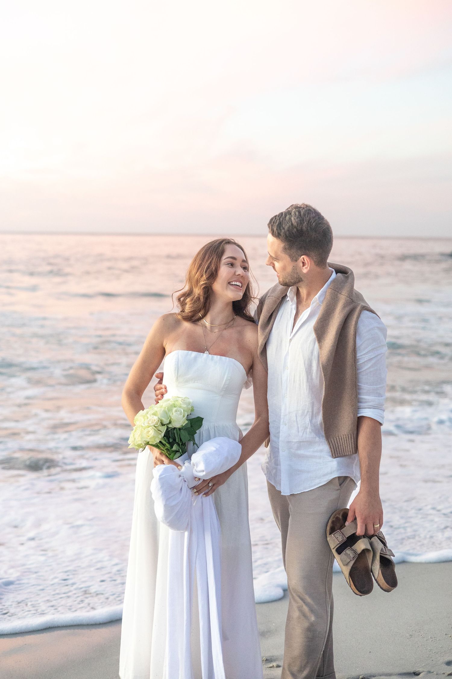 Romantic beach wedding scene at sunset with couple holding bouquet by ocean waves.