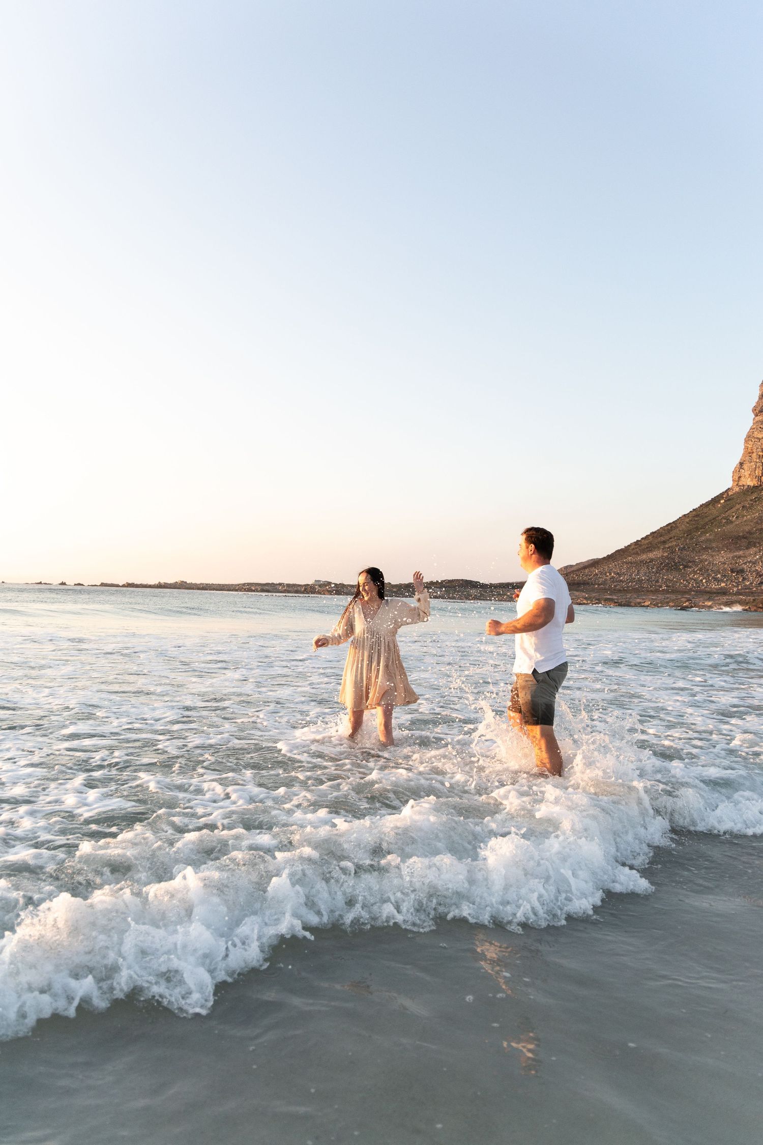 Couple playing in ocean waves at sunset with rocky cliffside in background.