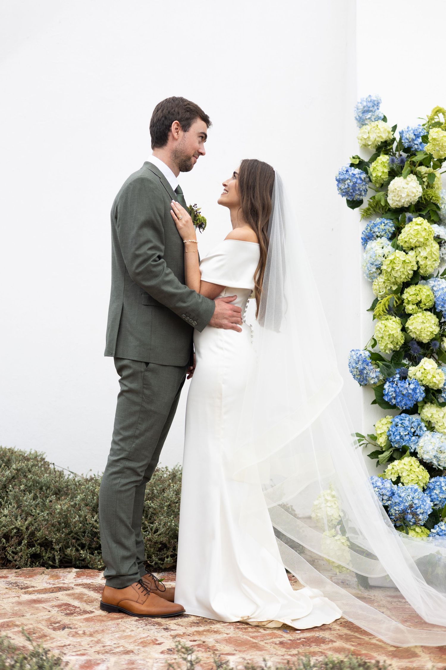 A couple in wedding attire poses against a backdrop of blue and green hydrangea flowers.