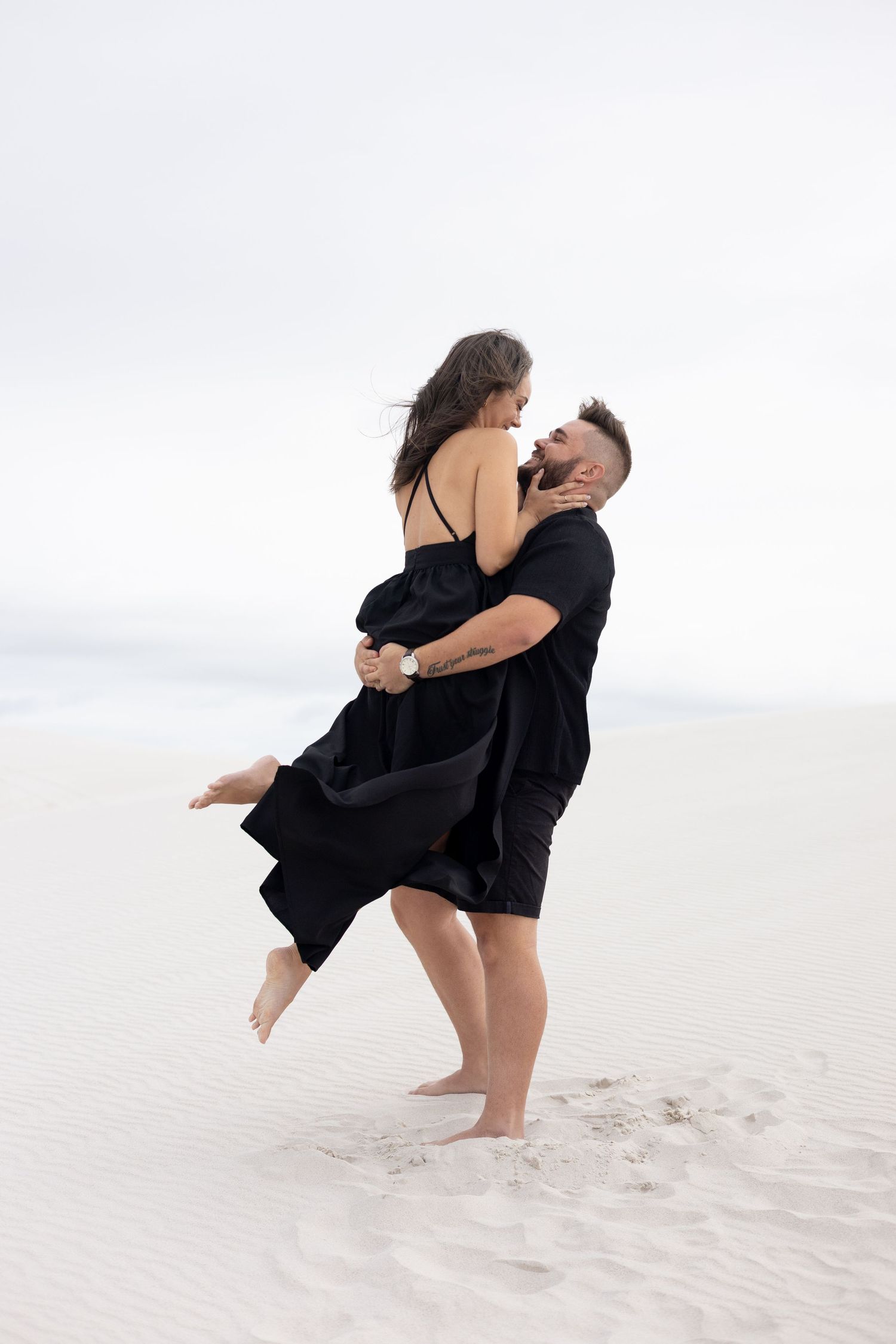Couple in black outfits embracing and dancing on white sand dunes under cloudy sky.