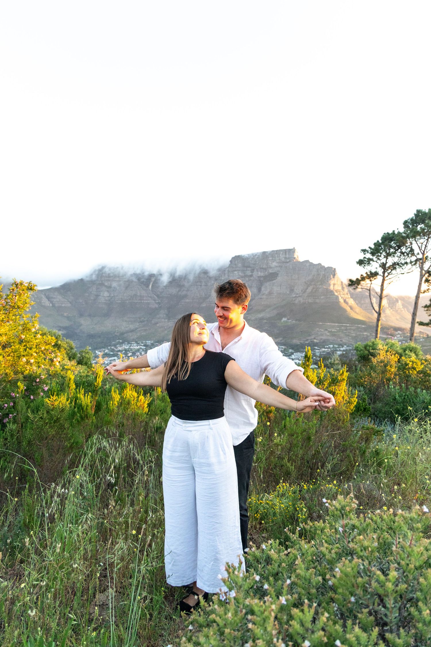 Couple poses playfully with arms outstretched among yellow wildflowers with mountain backdrop.