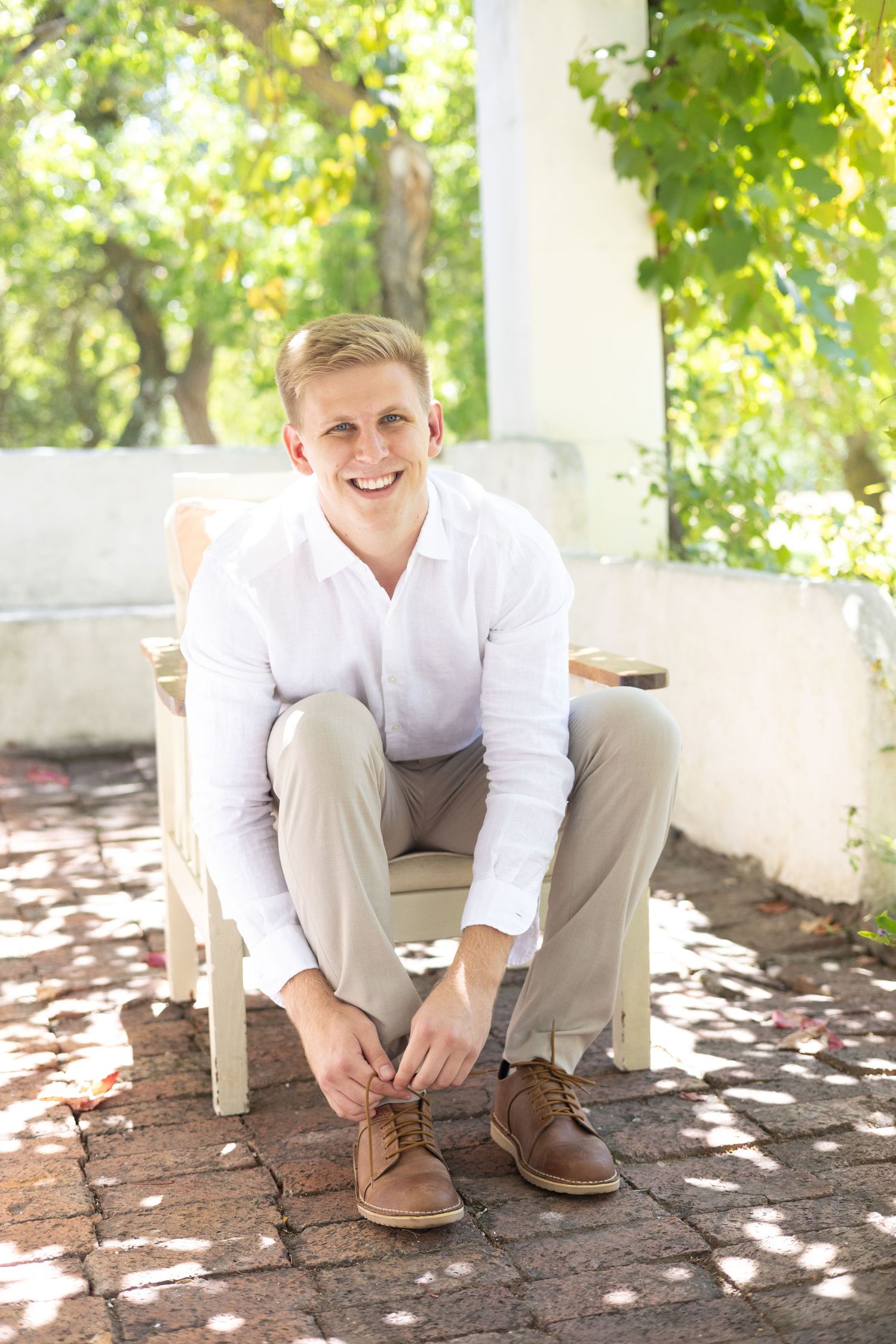 Person in white shirt and khaki pants sits on chair on outdoor patio with dappled sunlight.