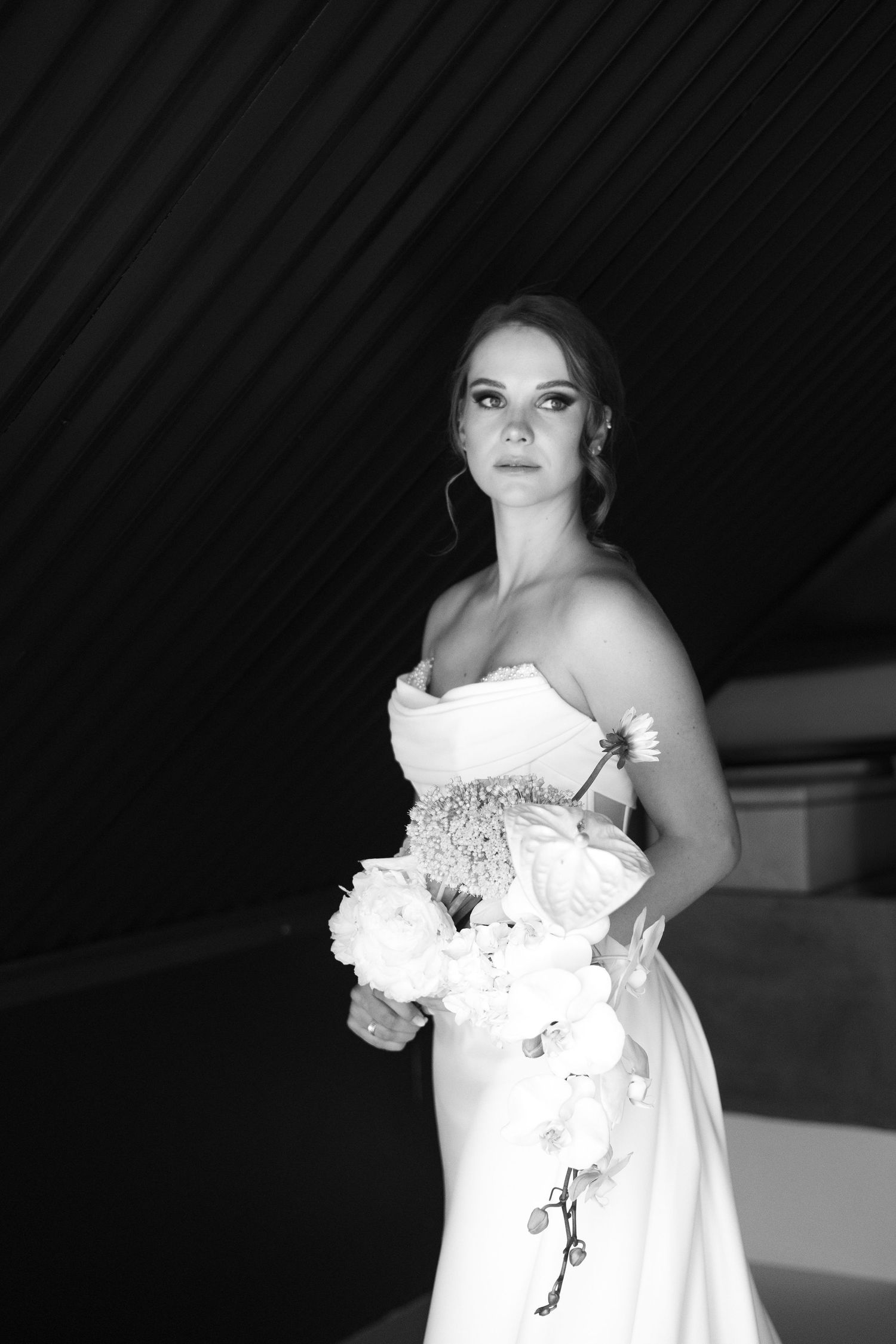 Black and white portrait of a bride in a strapless wedding gown holding a white bouquet against a dark background.