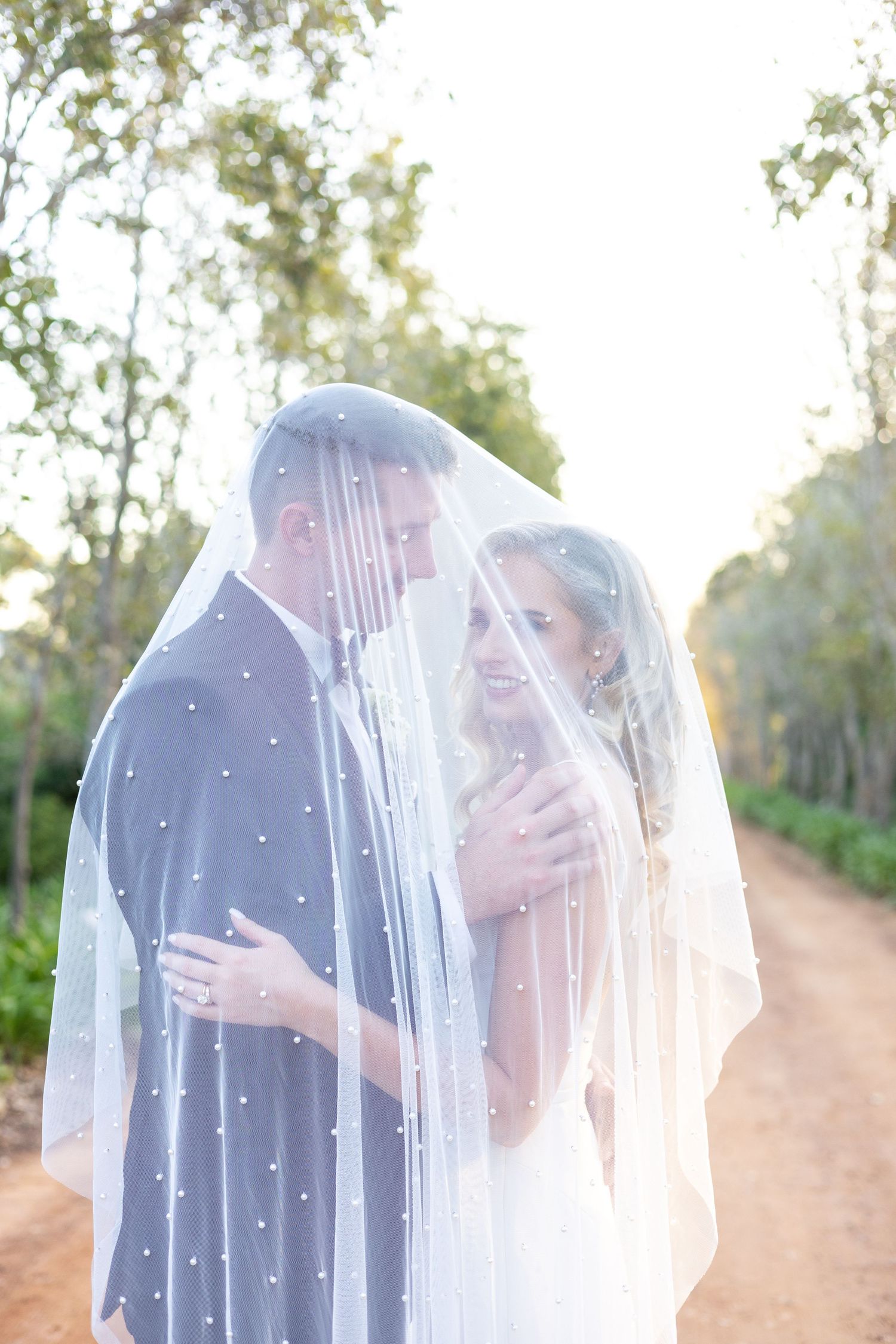 Romantic couple embraces under a sheer veil in an outdoor setting with soft sunlight filtering through trees.