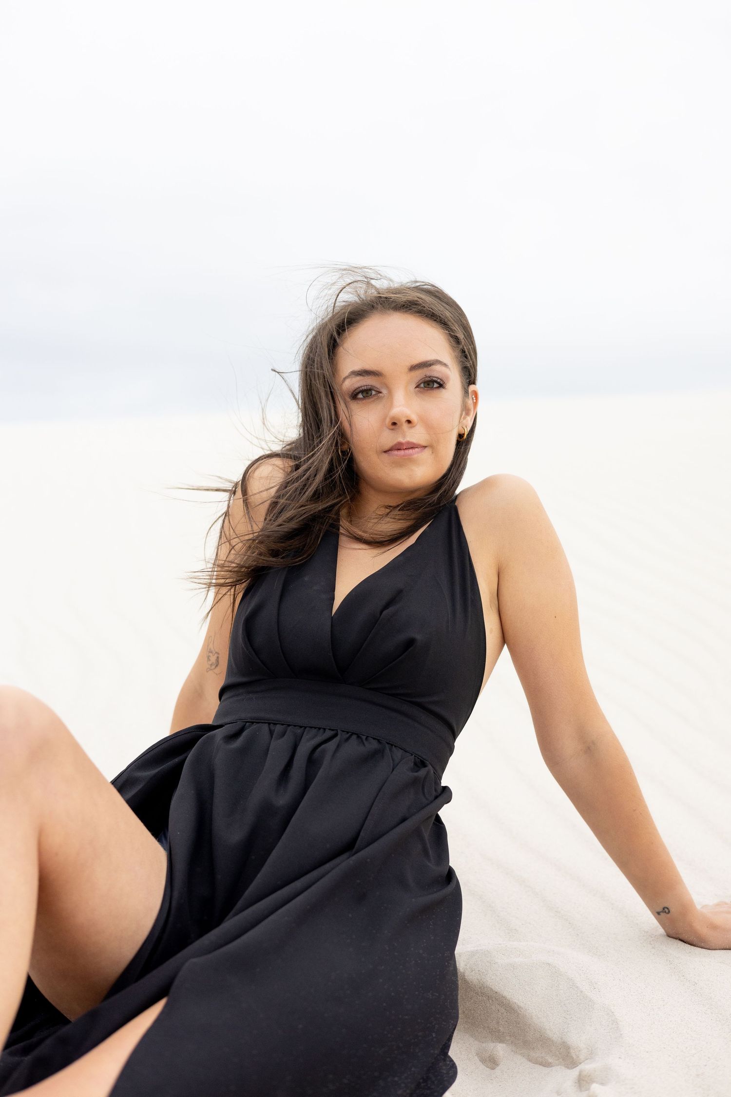 A woman in a black dress poses gracefully on white sand during a beach photoshoot.