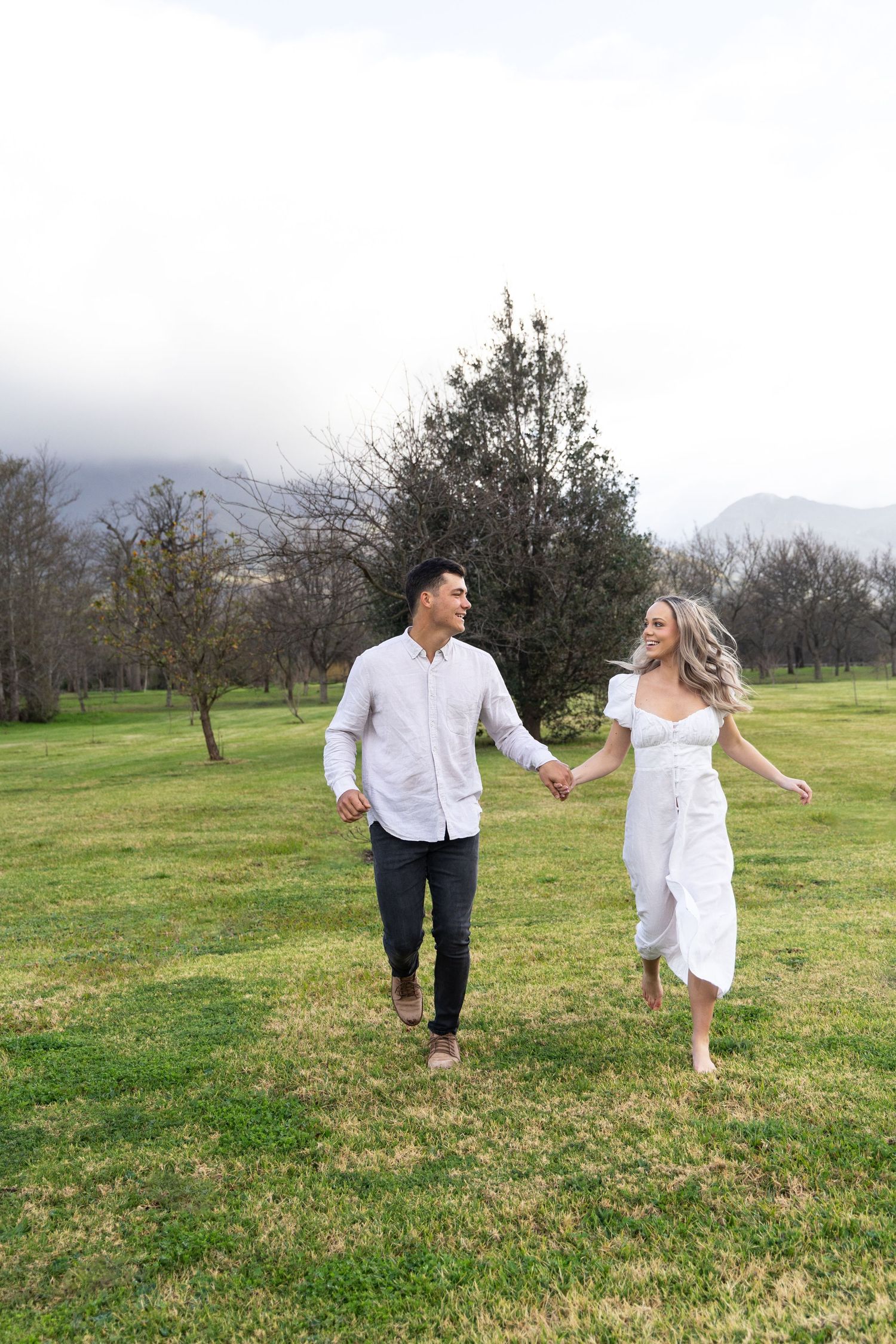 Couple in white outfits running hand-in-hand through green field with mountains in background.
