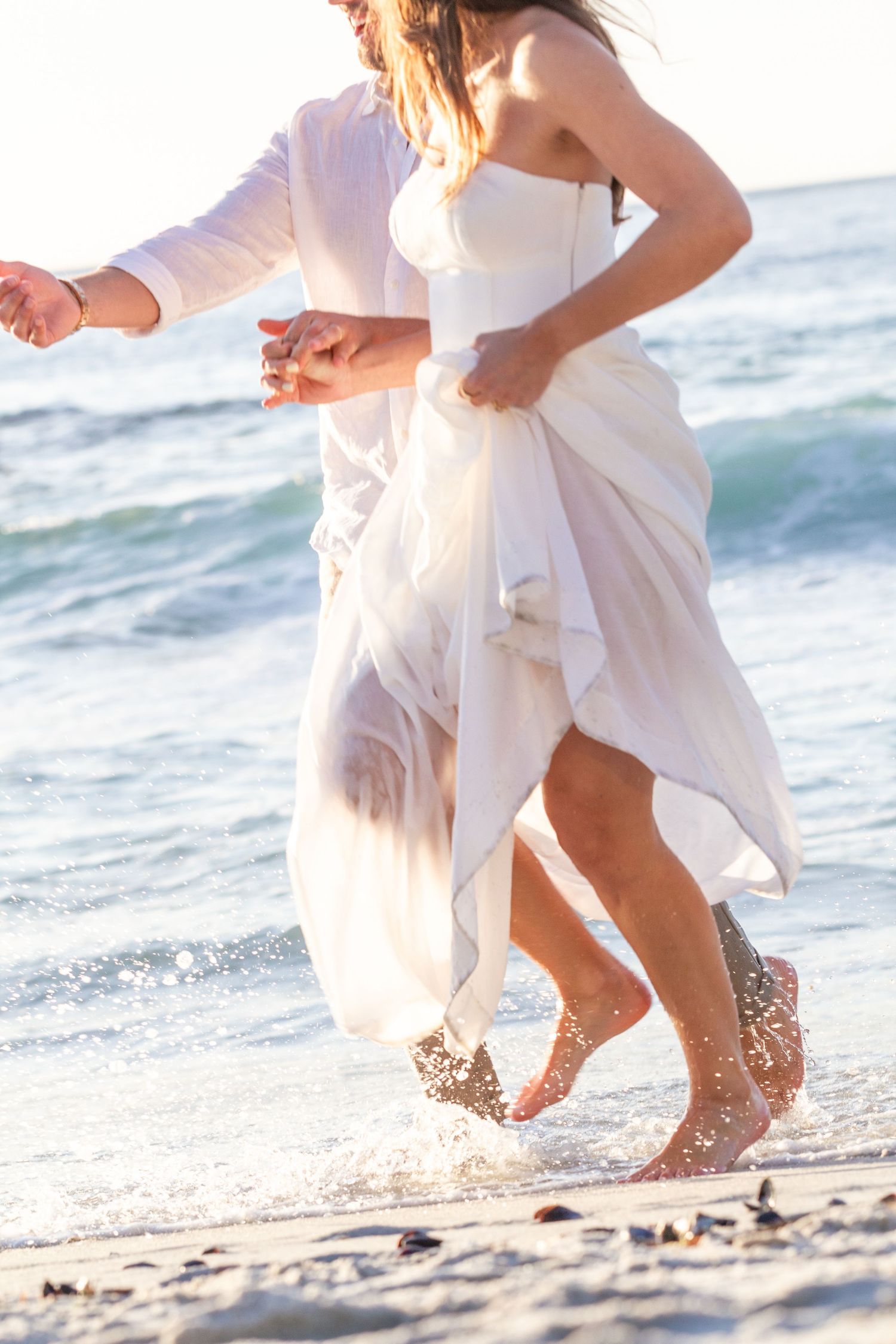 Couple dances playfully in white attire on sandy beach with waves in background.