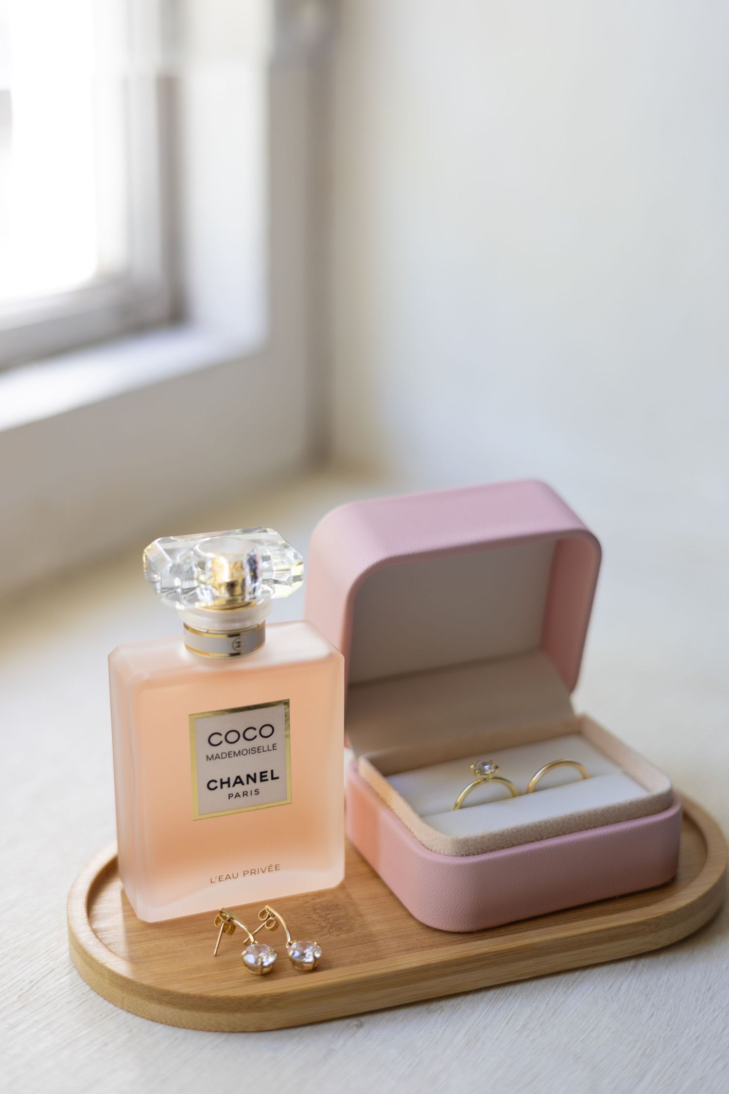 Chanel Coco Mademoiselle perfume bottle with jewelry displayed on wooden tray near window with pink ring box.