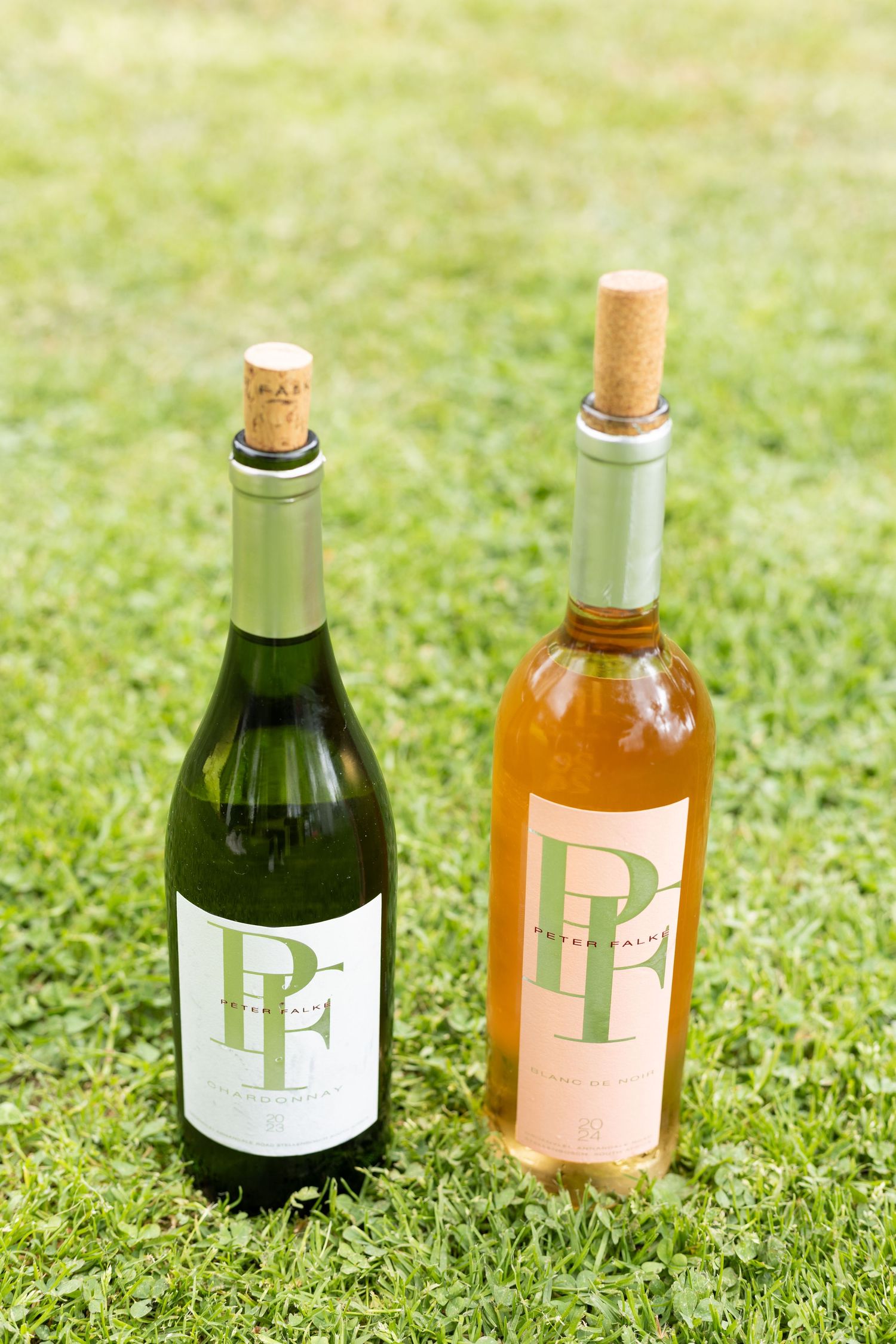 Two wine bottles with PP labels, one green glass and one rose colored, sitting on grass outdoors.