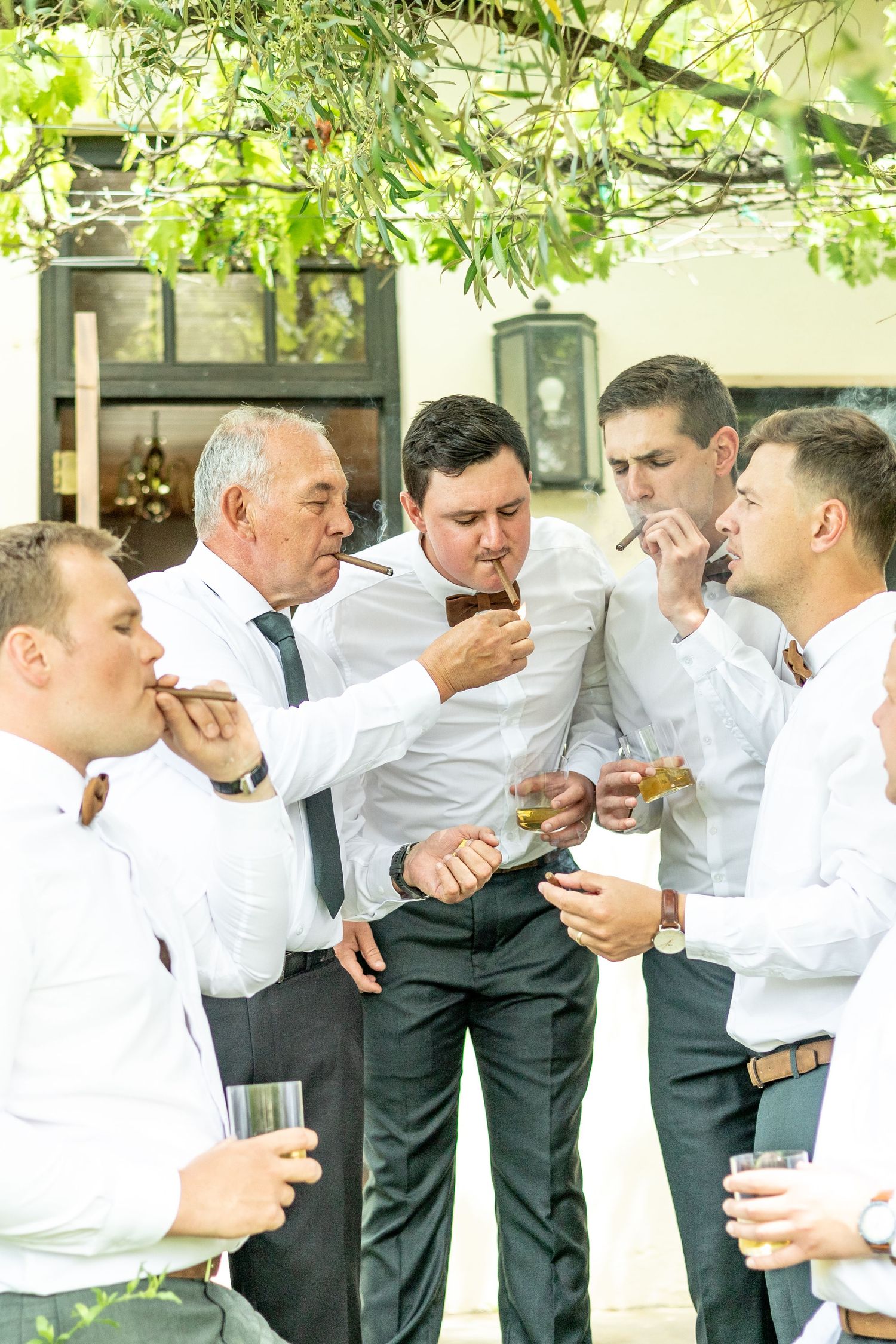 A group of men in white shirts and dark pants gather together sharing drinks and cigars outdoors.