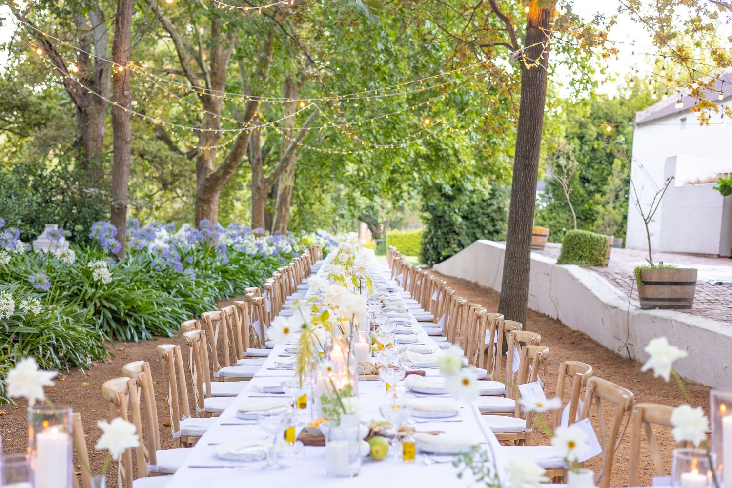 A long outdoor wedding reception table with white linens and wooden chairs surrounded by lush garden landscaping.