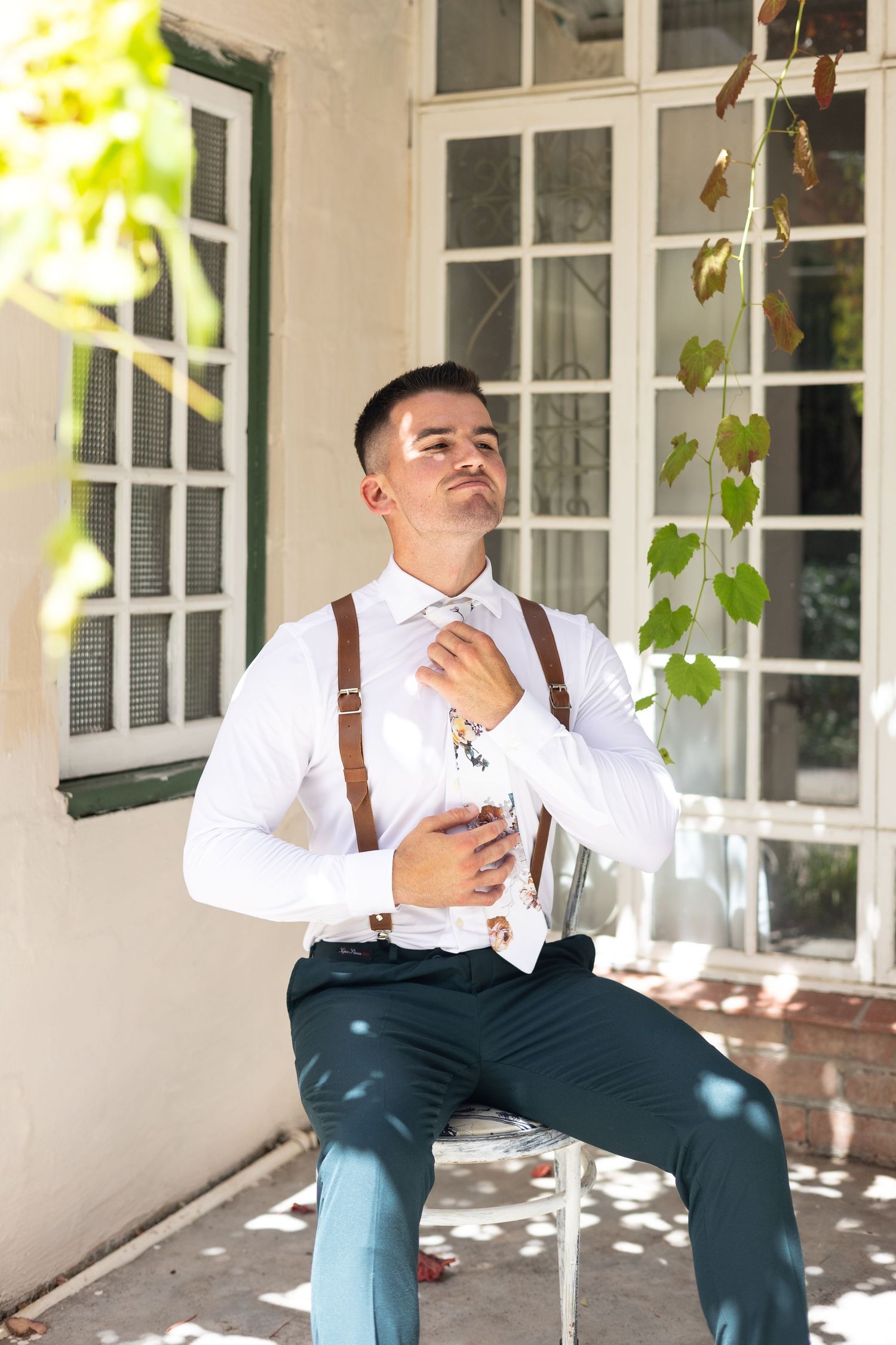 A person in formal attire with brown suspenders and white shirt sits on a chair outdoors near white windows and vines.