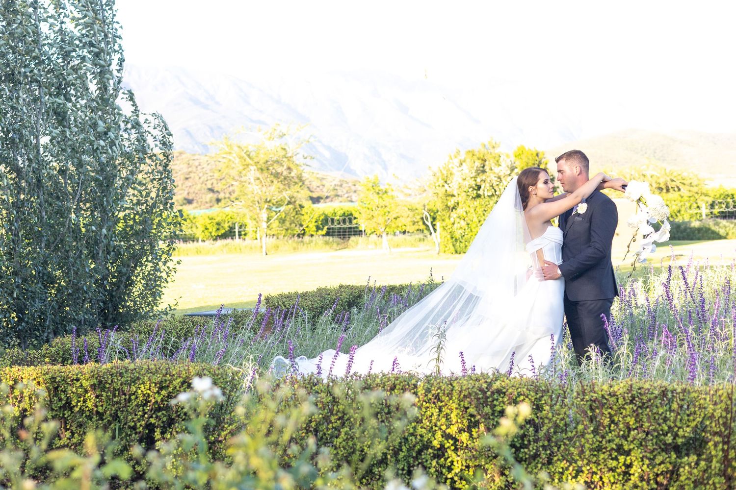 A couple shares a romantic moment in a garden landscape with wildflowers and trees bathed in golden sunlight.