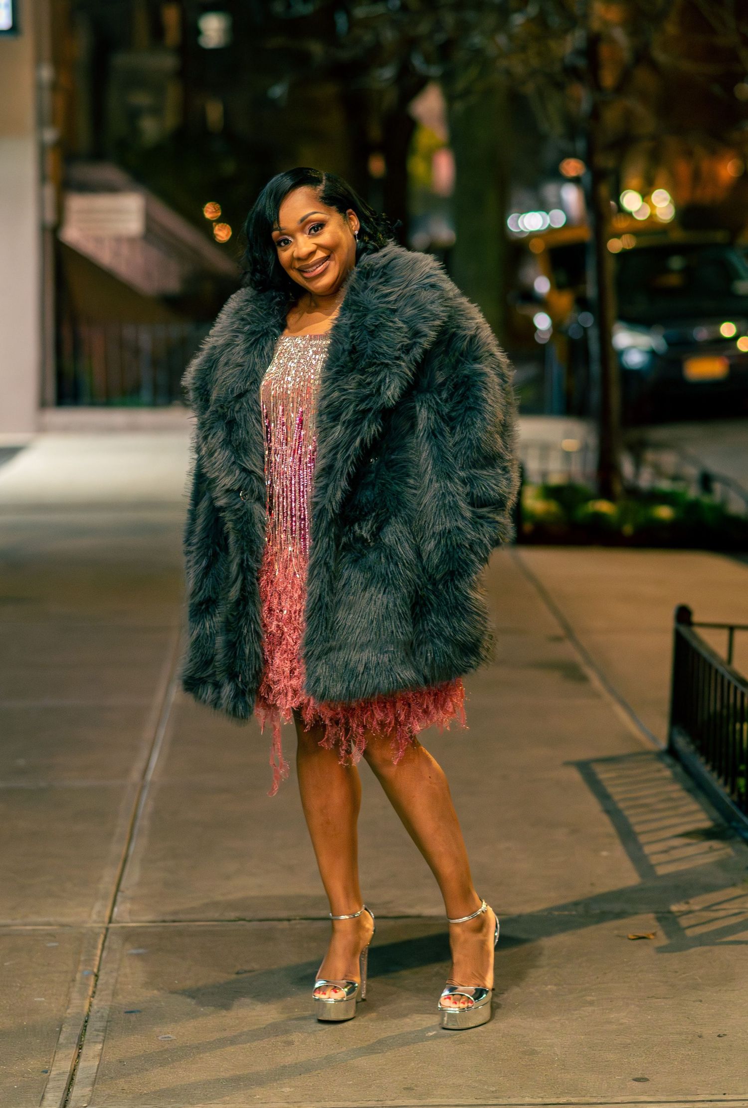 Stylish outfit featuring grey fur coat and sparkly dress captured on city sidewalk at night.