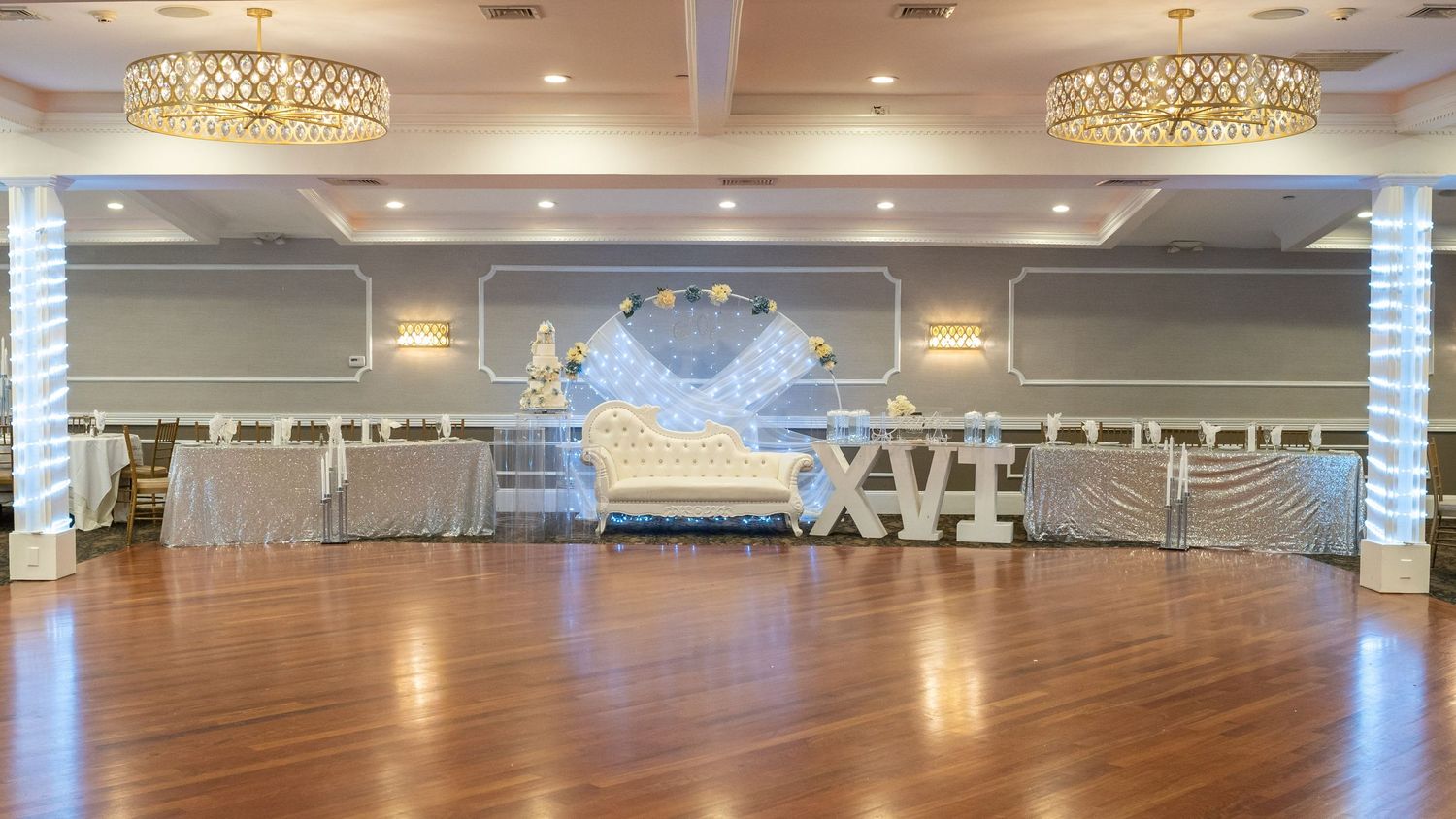 Elegant wedding reception venue with white throne seating and chandelier lighting on hardwood floor.