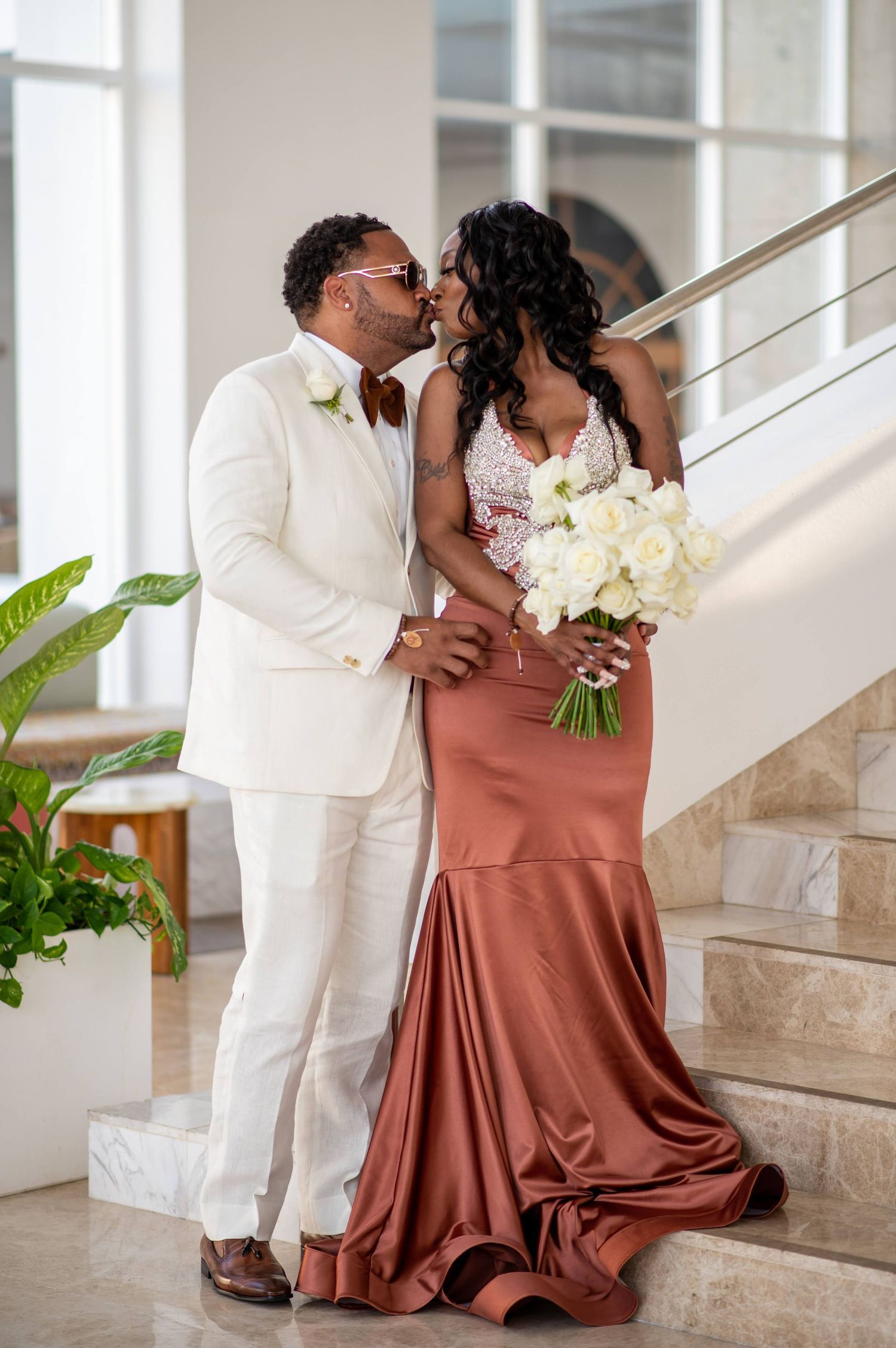 Romantic couple in white and rose formal attire embracing on elegant staircase with bouquet.