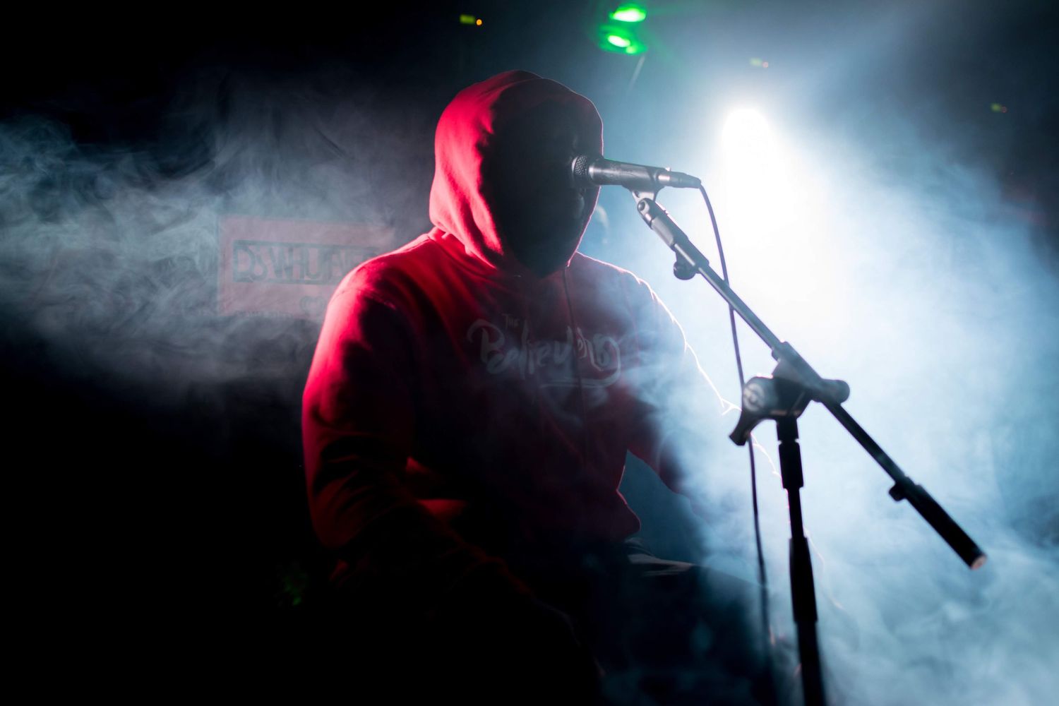 A silhouetted performer in a hoodie sings into a microphone surrounded by atmospheric stage fog.