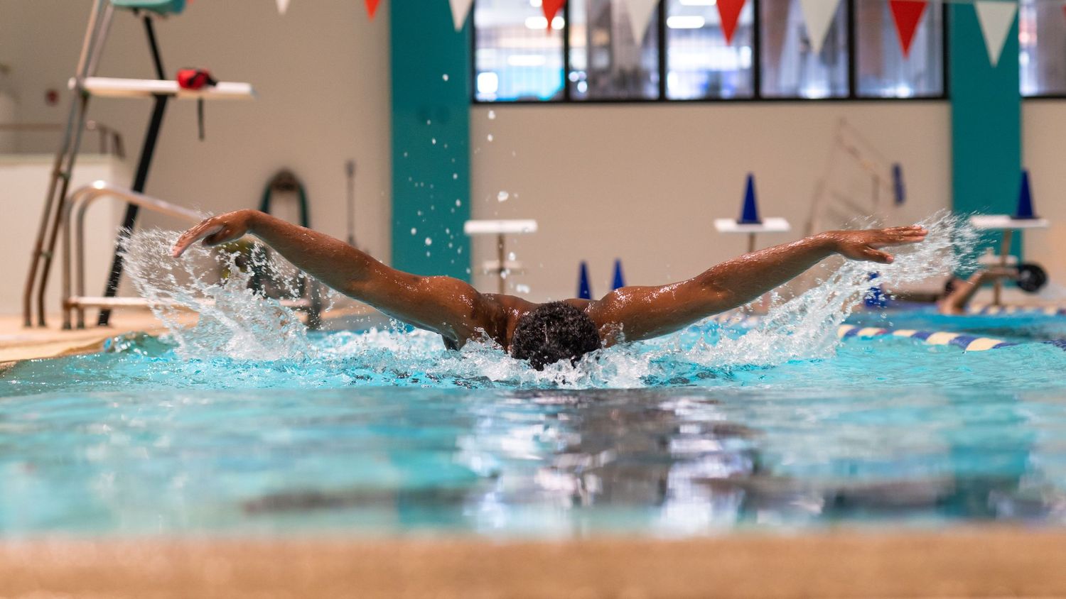 Swimmer performing butterfly stroke in indoor pool with water splashing around.