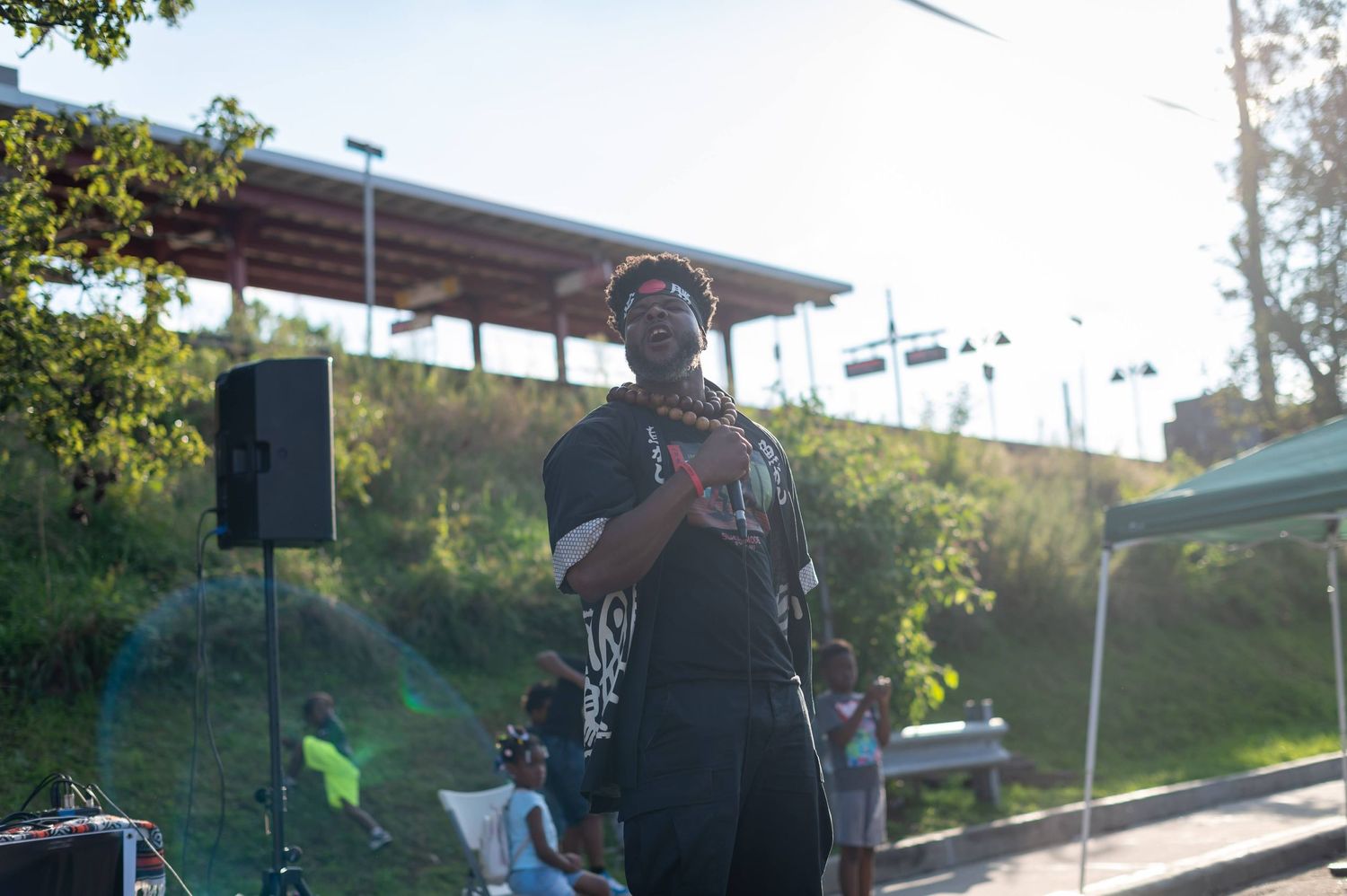 A performer delivers an energetic outdoor performance in front of speakers and greenery.