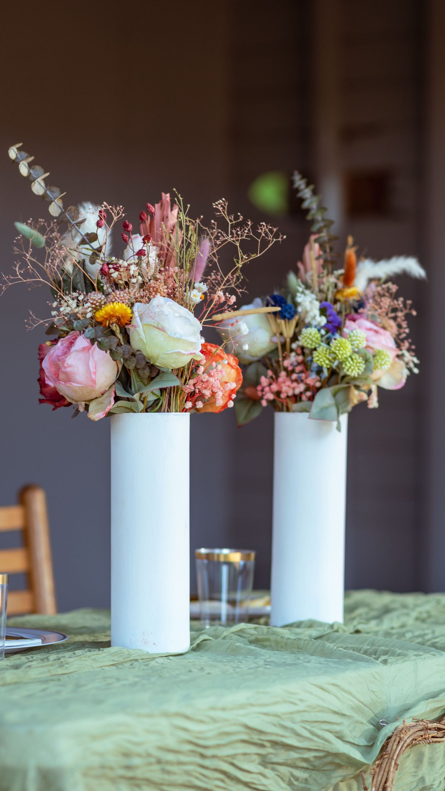 Two elegant white cylindrical vases filled with colorful spring flowers sit atop a mint green tablecloth.