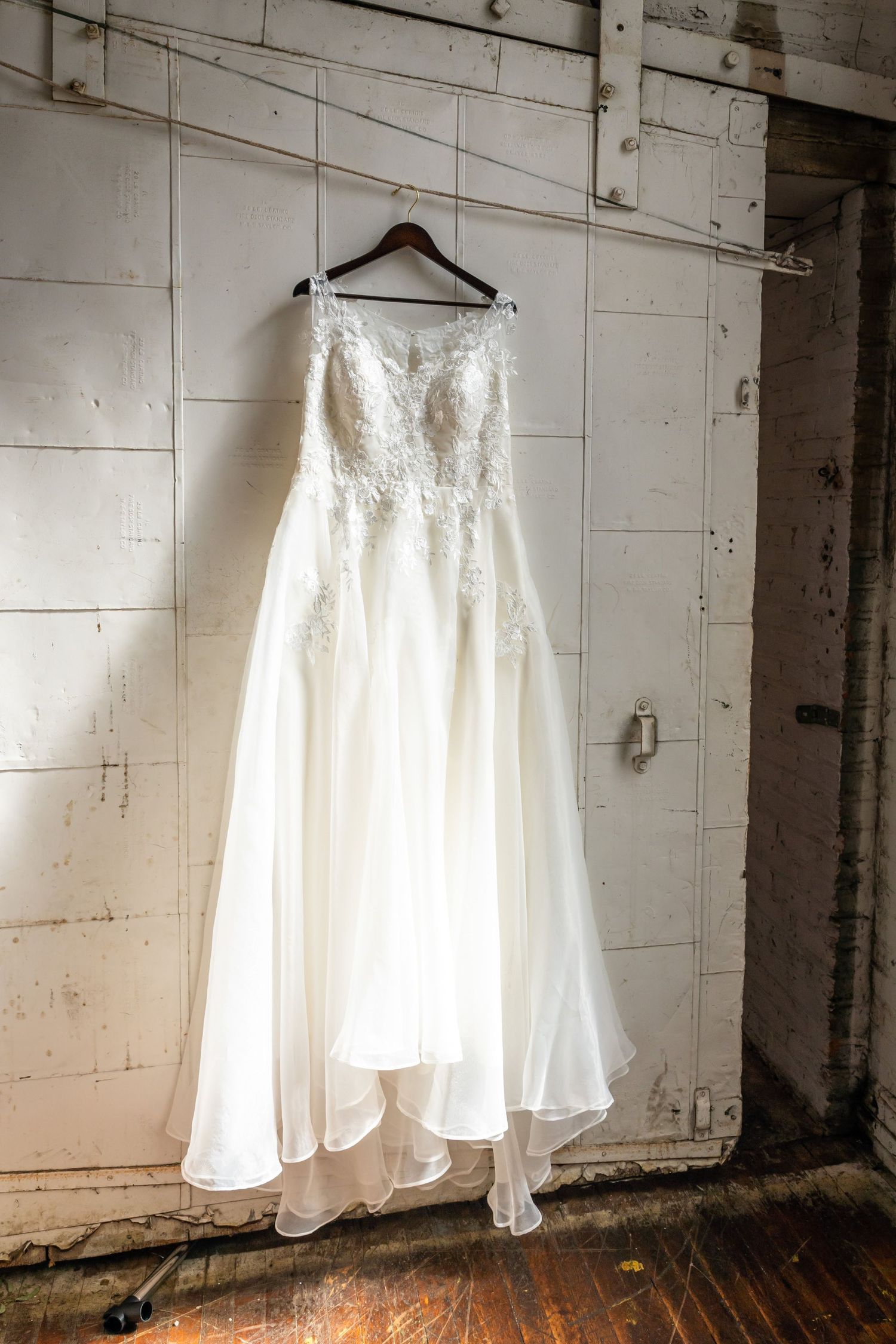 A white lace wedding dress hanging on display against a rustic industrial wall with natural lighting.