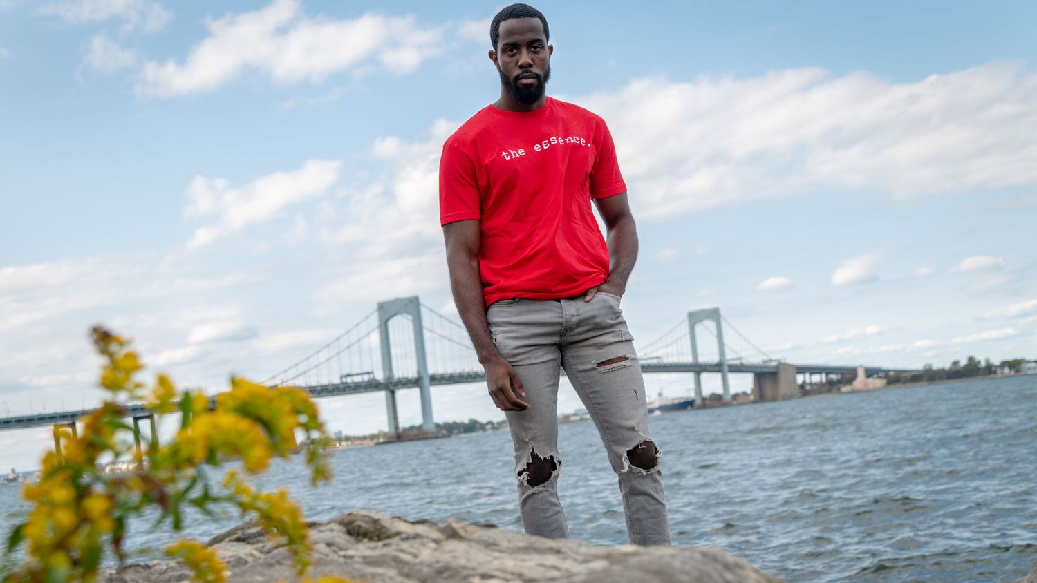A person in a red shirt stands near the waterfront with a bridge visible in the background.