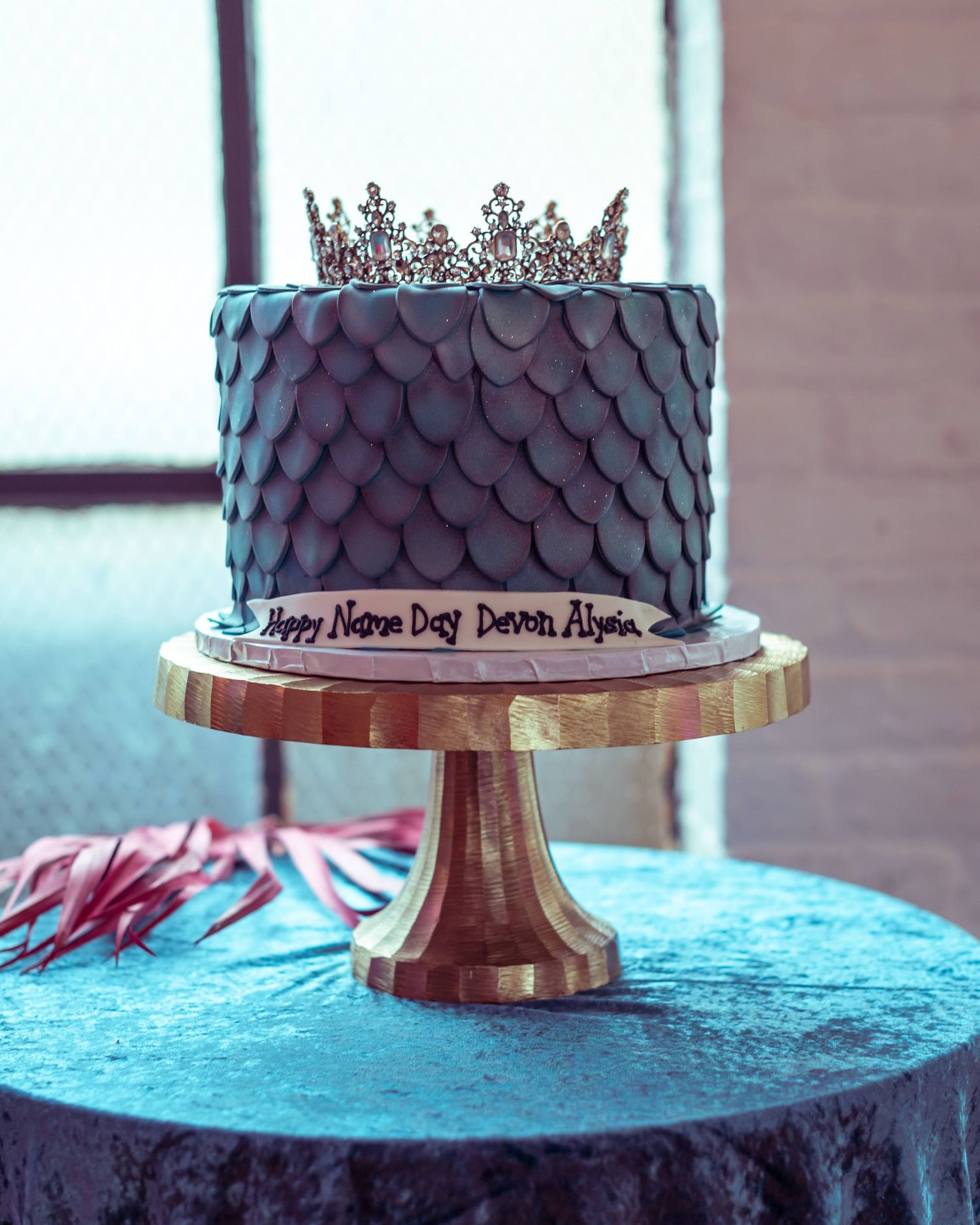 Dark grey textured cake with metallic crown topping on a gold cake stand against a turquoise backdrop.