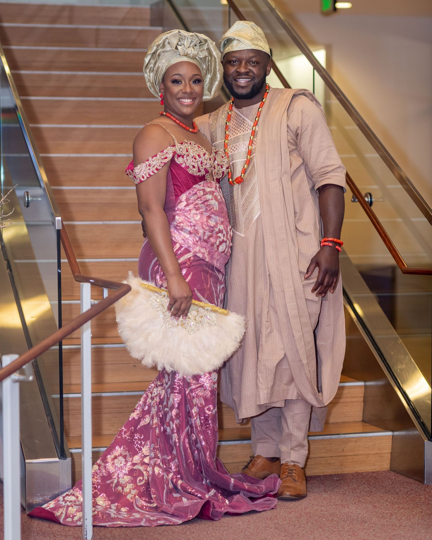 A couple in traditional Nigerian wedding attire posing on stairs with a pink gown and cream agbada.