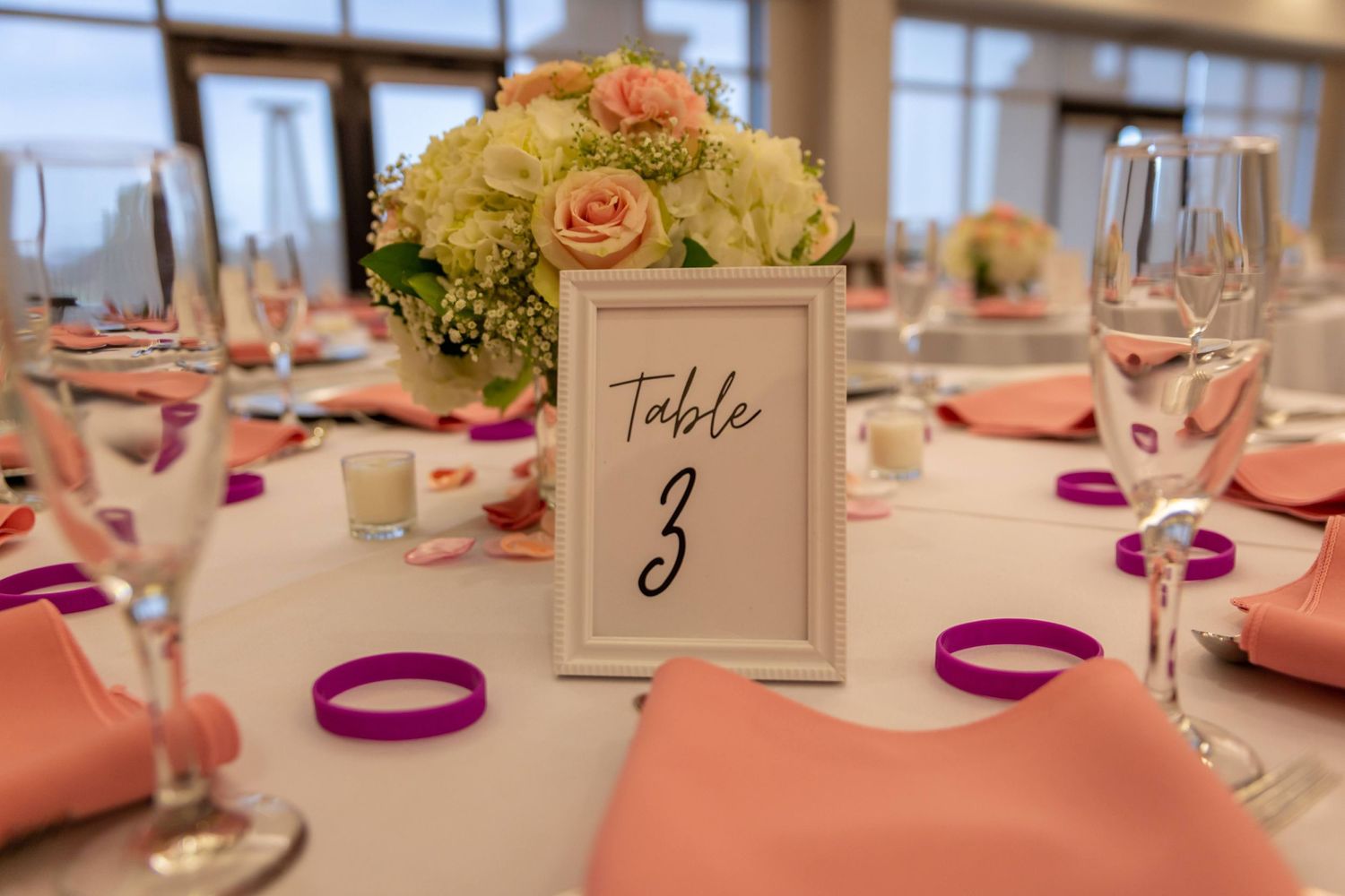 An elegant wedding reception table setting with coral napkins, floral centerpiece and table number three.