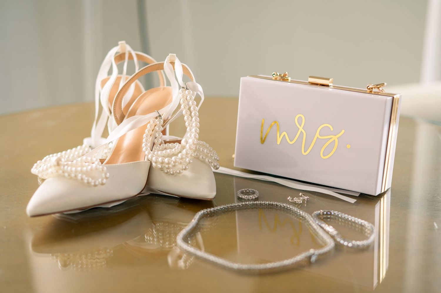 Bridal accessories flat lay with pearl heels, Mrs. clutch purse, diamond rings, and pearl necklace on gold surface.