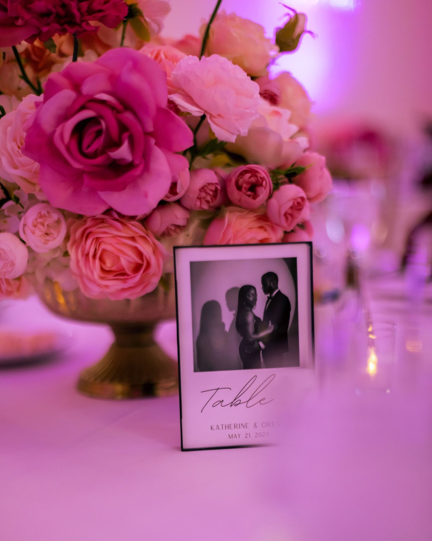 A pink floral centerpiece with roses surrounds a framed table number at a wedding reception.
