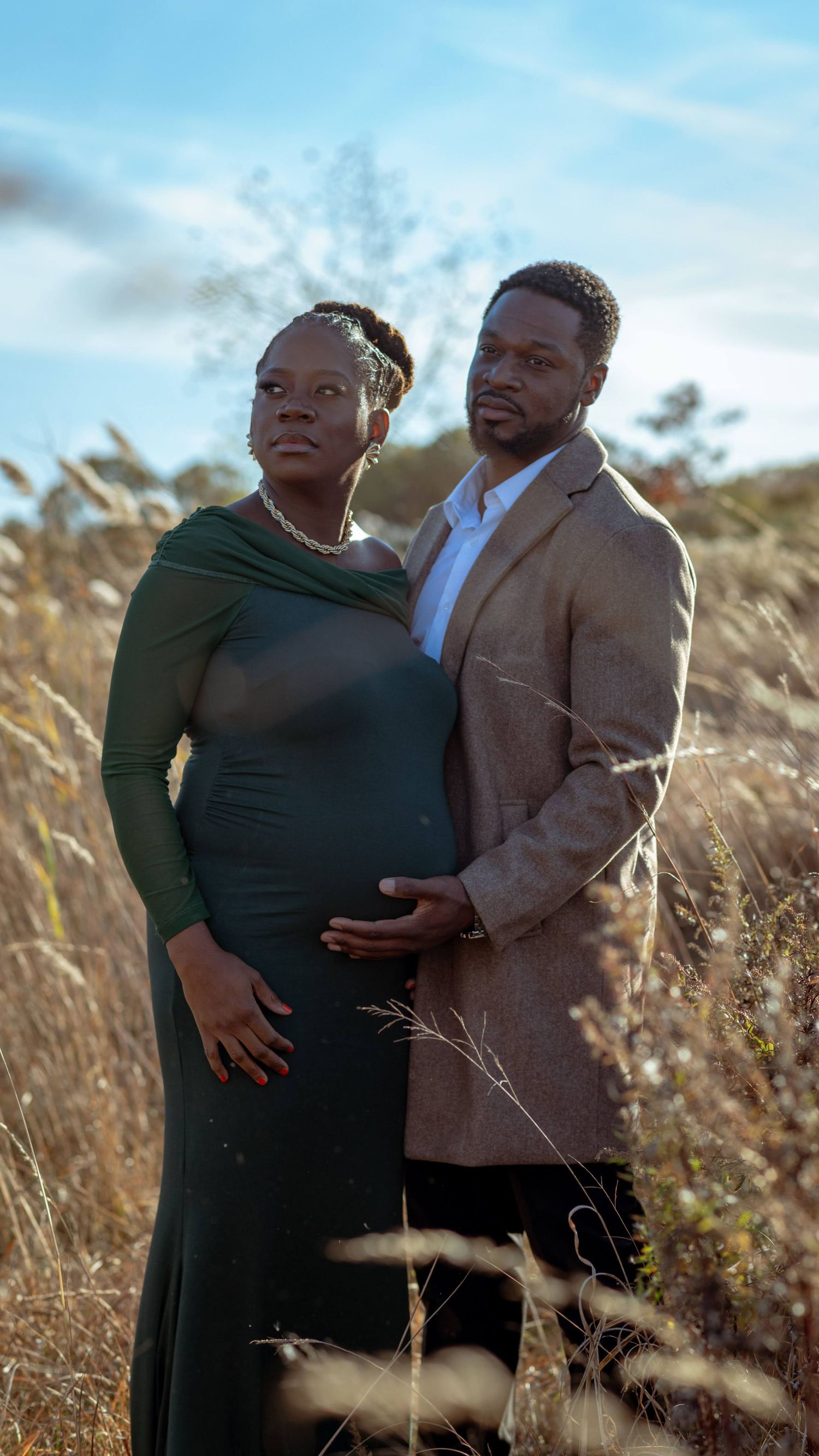 A maternity photoshoot in a field of tall grass with warm sunlight and a blue sky in the background.
