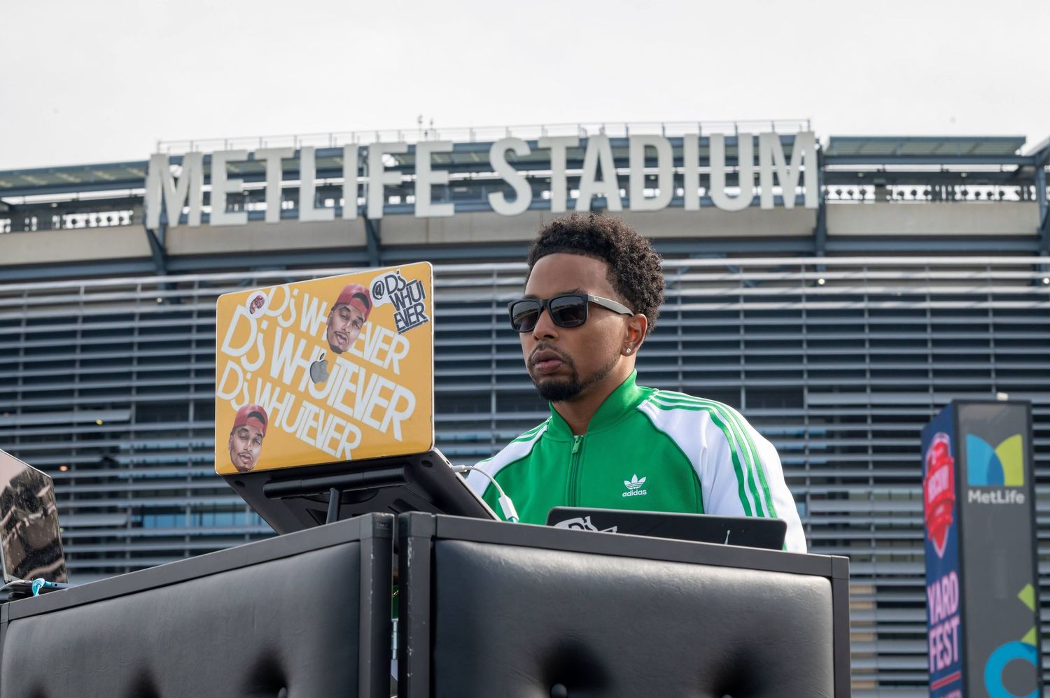 DJ performs at outdoor stadium event wearing green jacket and sunglasses.