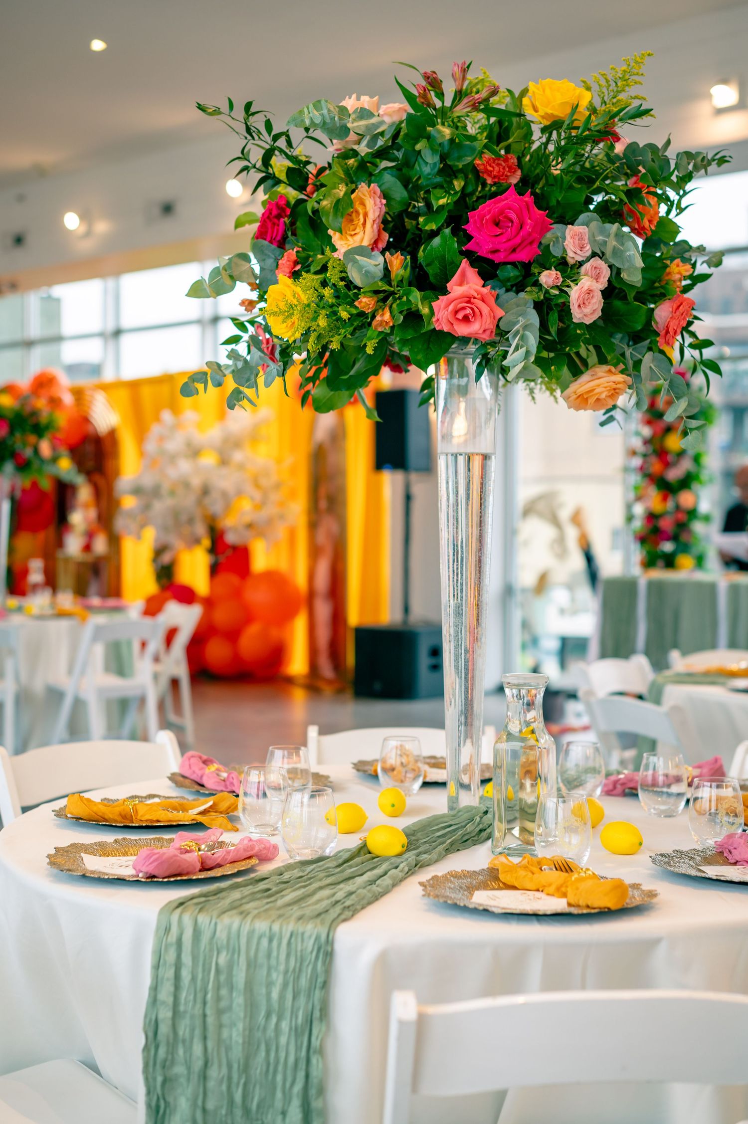 Elegant event table with tall floral centerpiece featuring colorful roses, greenery, white chairs, and sage green table runner.