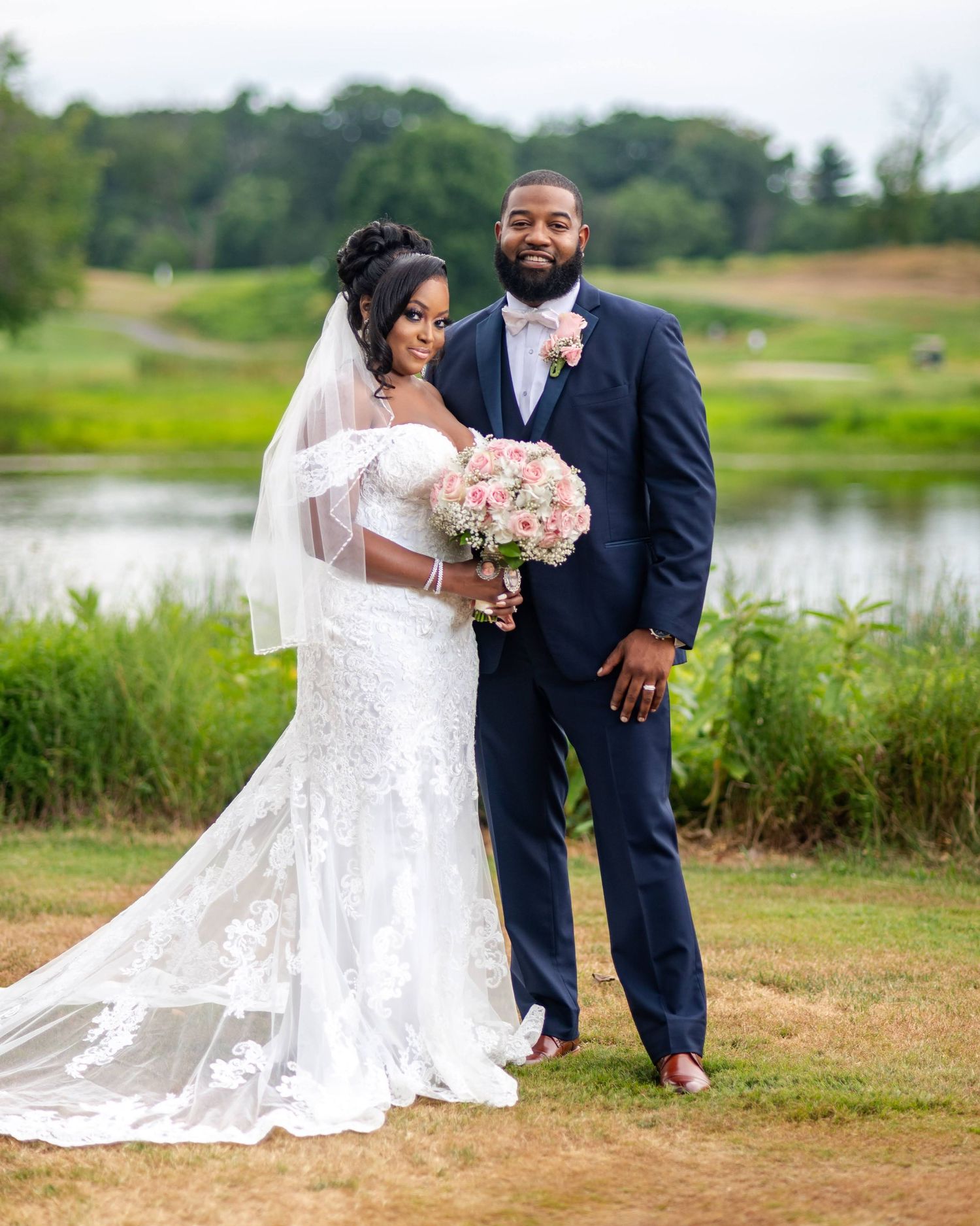 Wedding couple poses by lake with lace gown and pink bouquet.