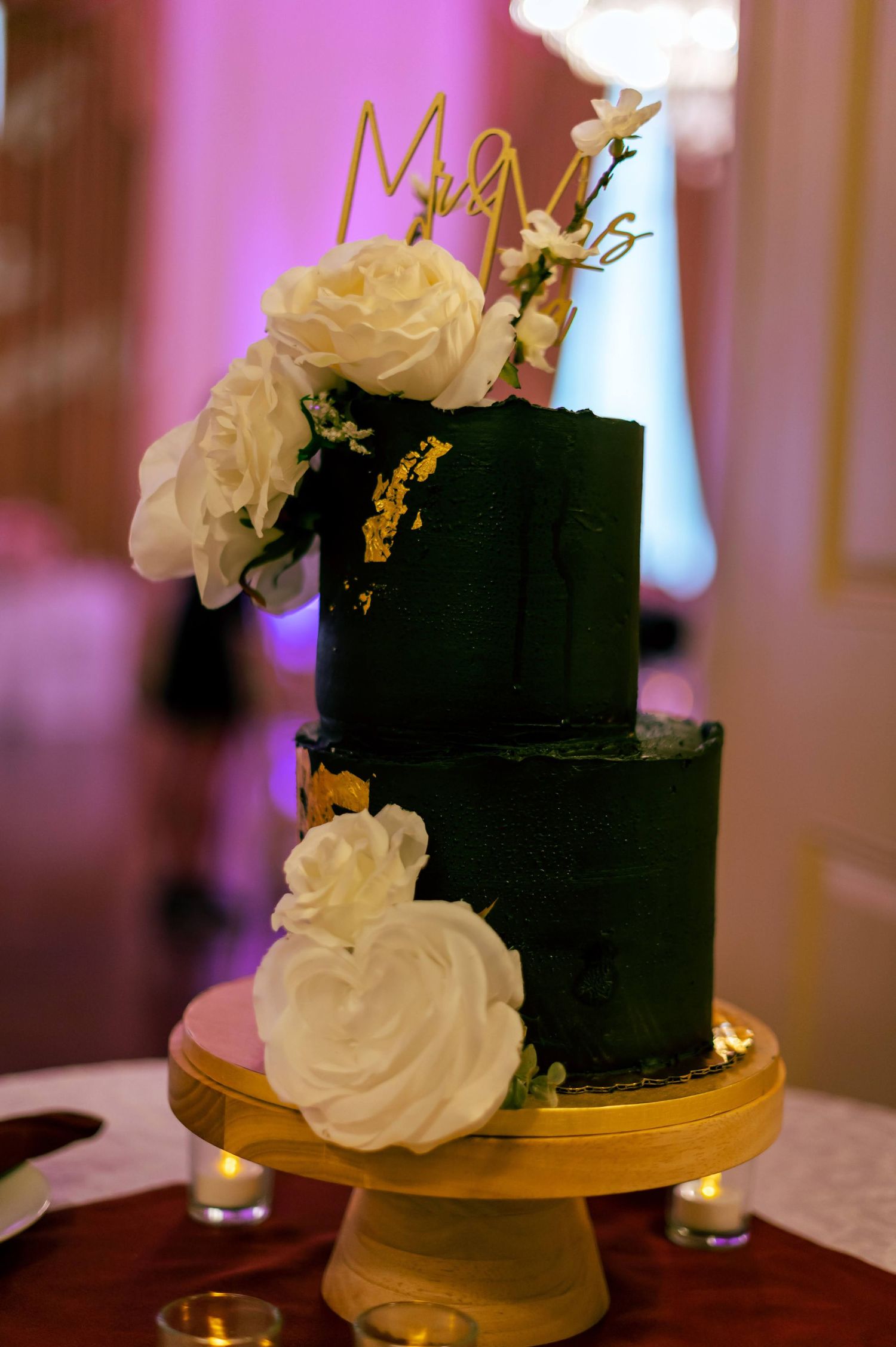 An elegant black wedding cake decorated with white roses and gold accents sits on a gold cake stand.