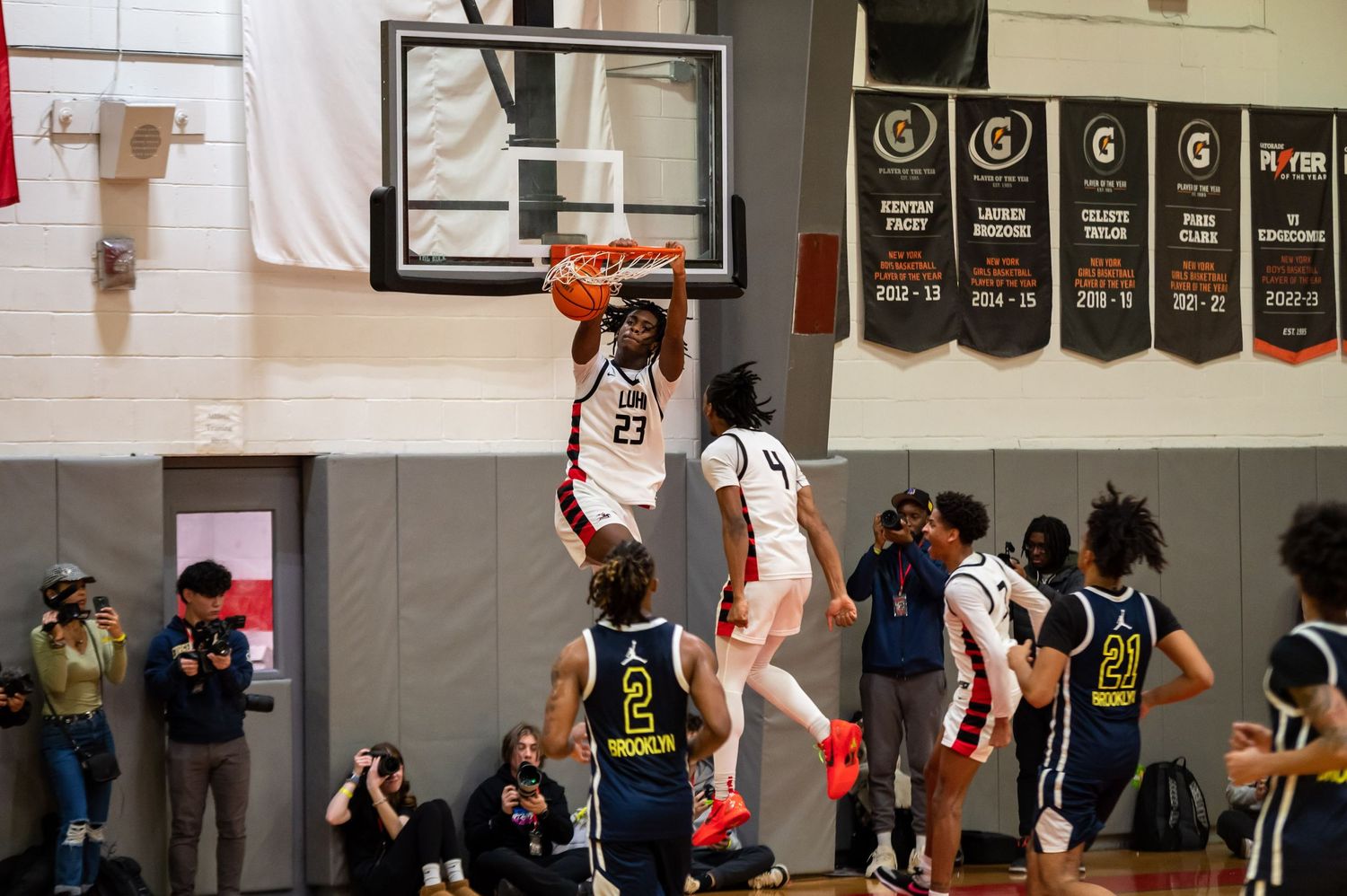 Basketball player jumps high for a dramatic layup during an indoor game with championship banners visible.