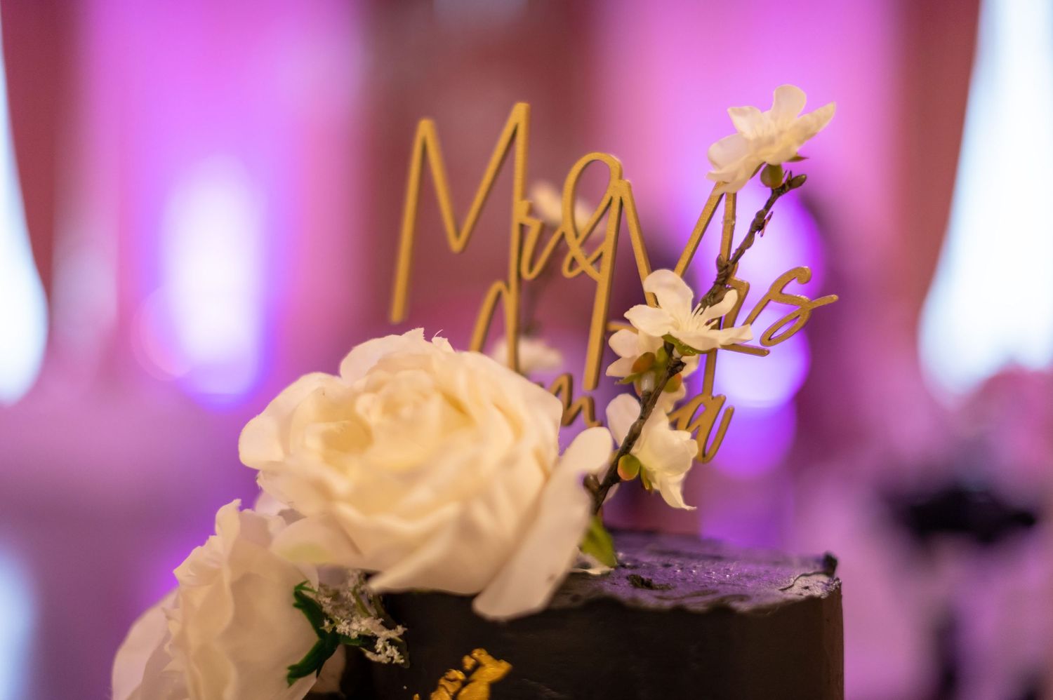 A black and gold wedding cake topper detail with white roses against pink ambient lighting.