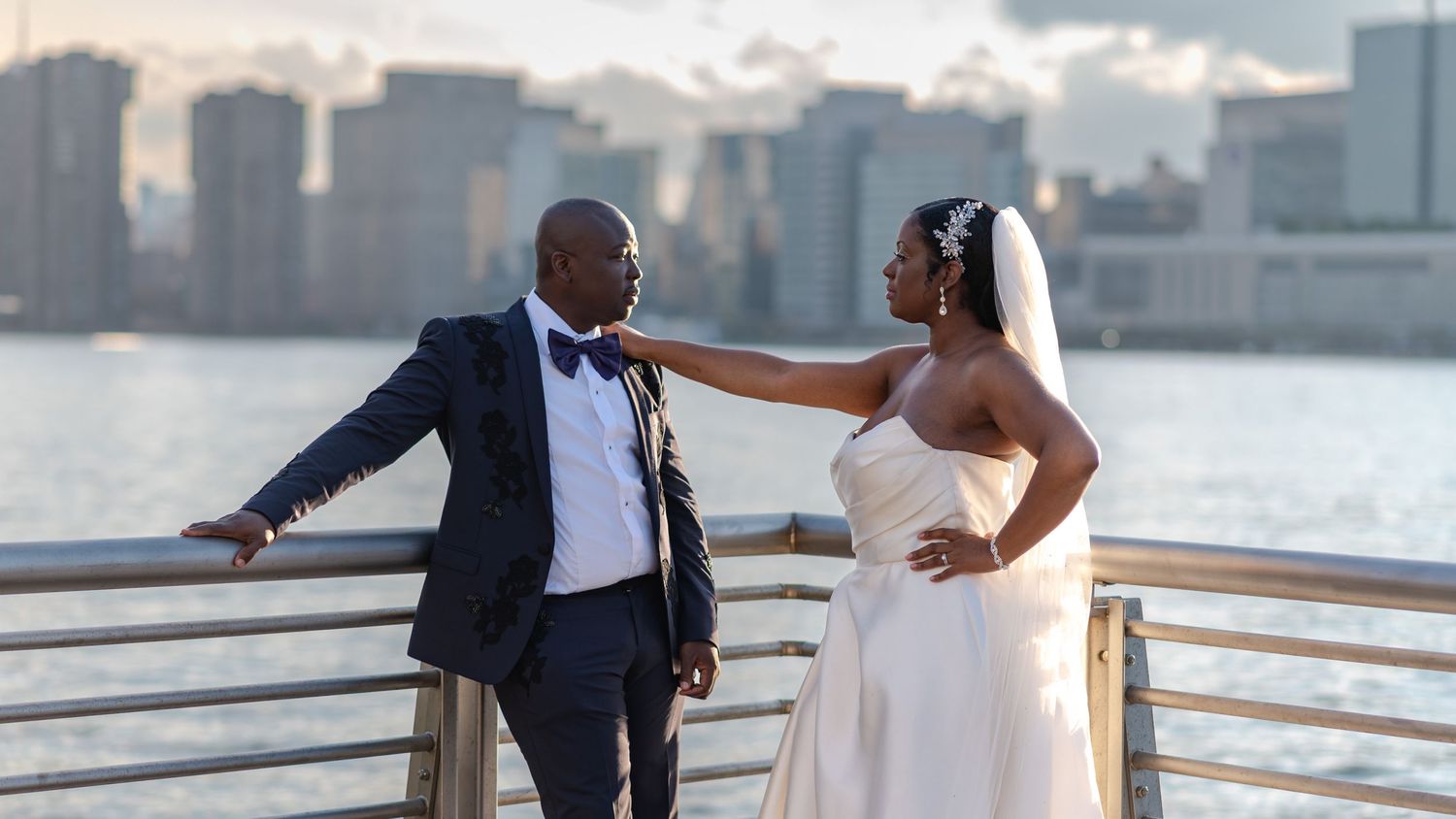 Couple in wedding attire posing on waterfront with city skyline in background.