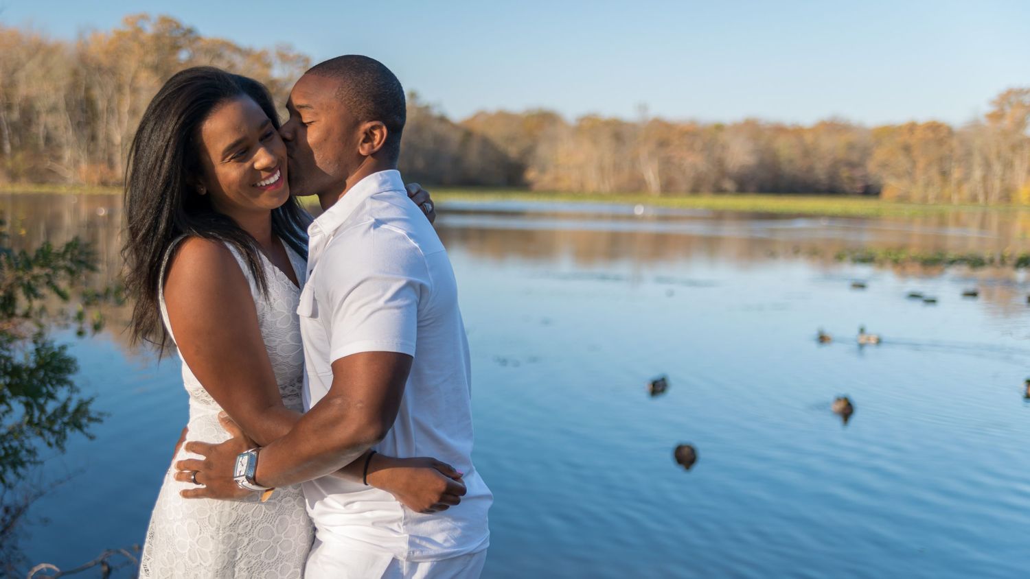 A romantic couple embraces by a serene lake during golden hour in casual white outfits.