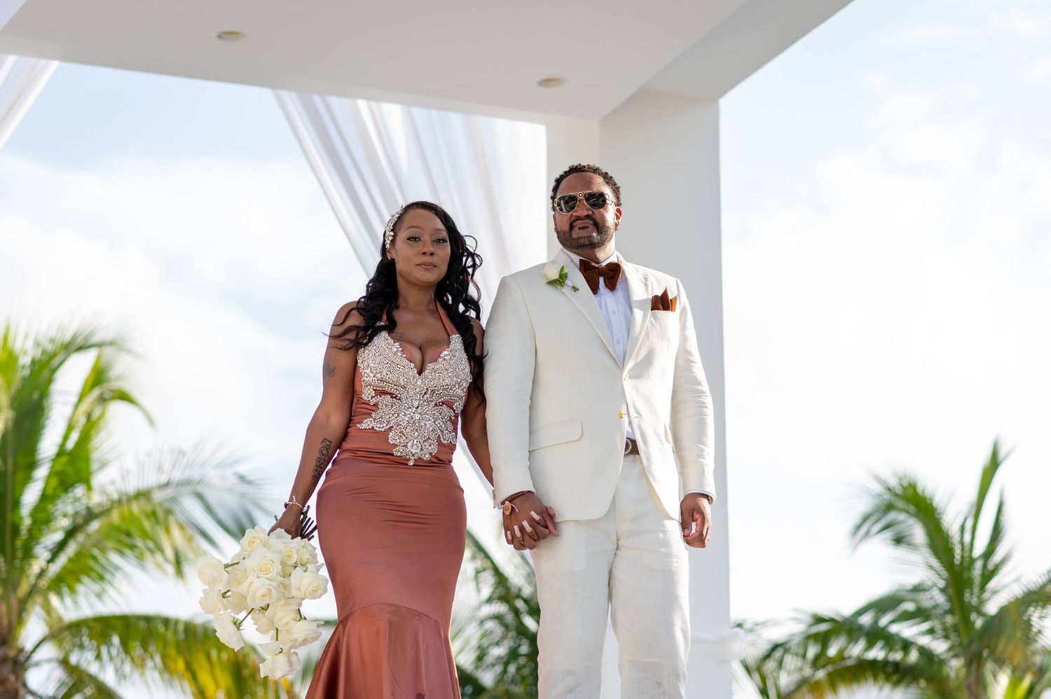 A couple in wedding attire standing on a balcony with palm trees in the background.