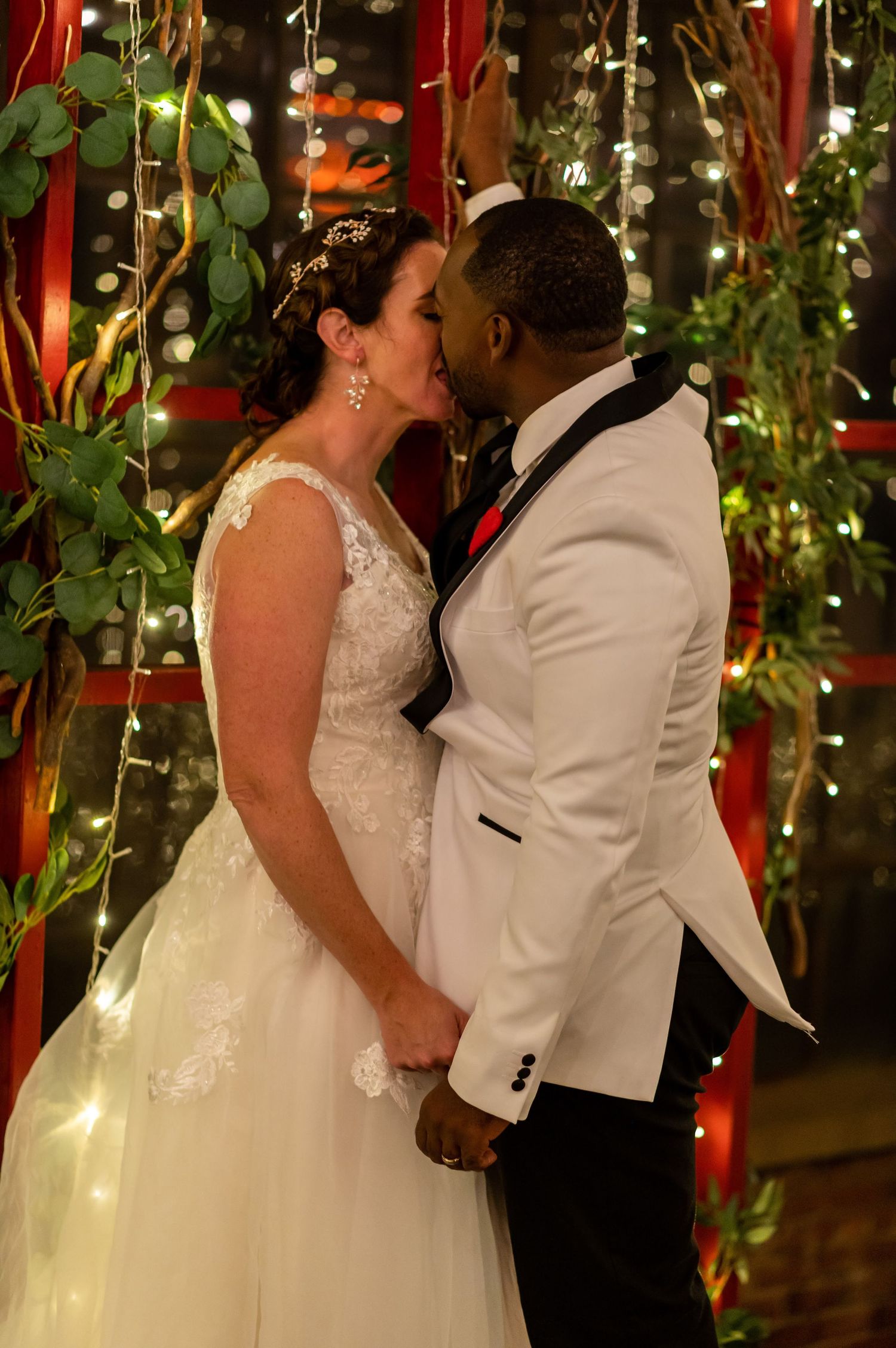 Romantic kiss between newlyweds under twinkling lights and greenery decorations.