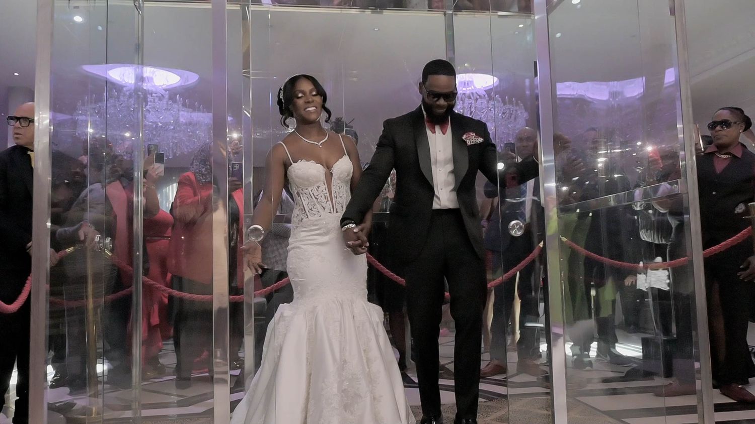 Newlyweds make grand exit through modern glass corridor with purple lighting.