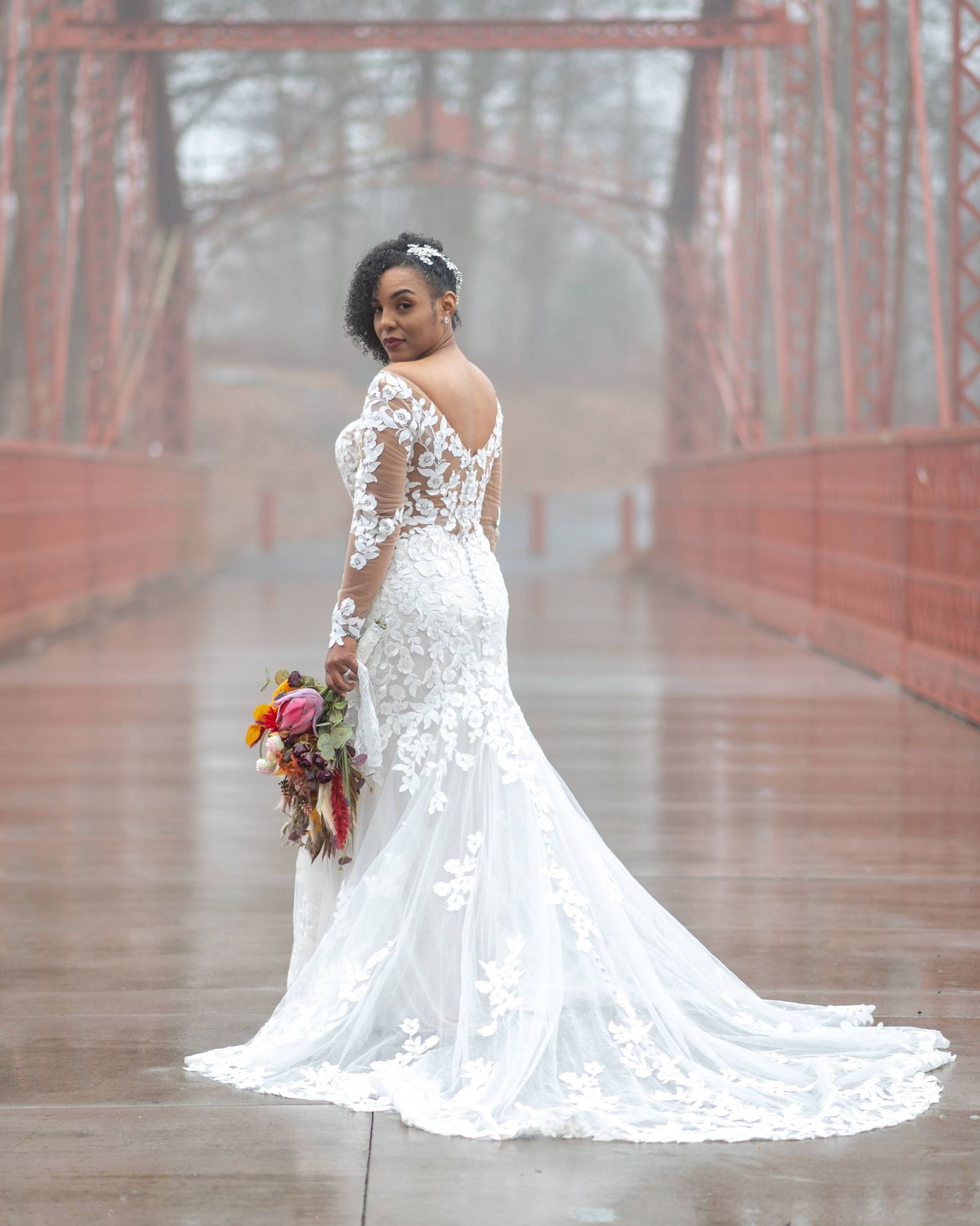 A bride in a lace wedding gown poses on a bridge holding a colorful bouquet.