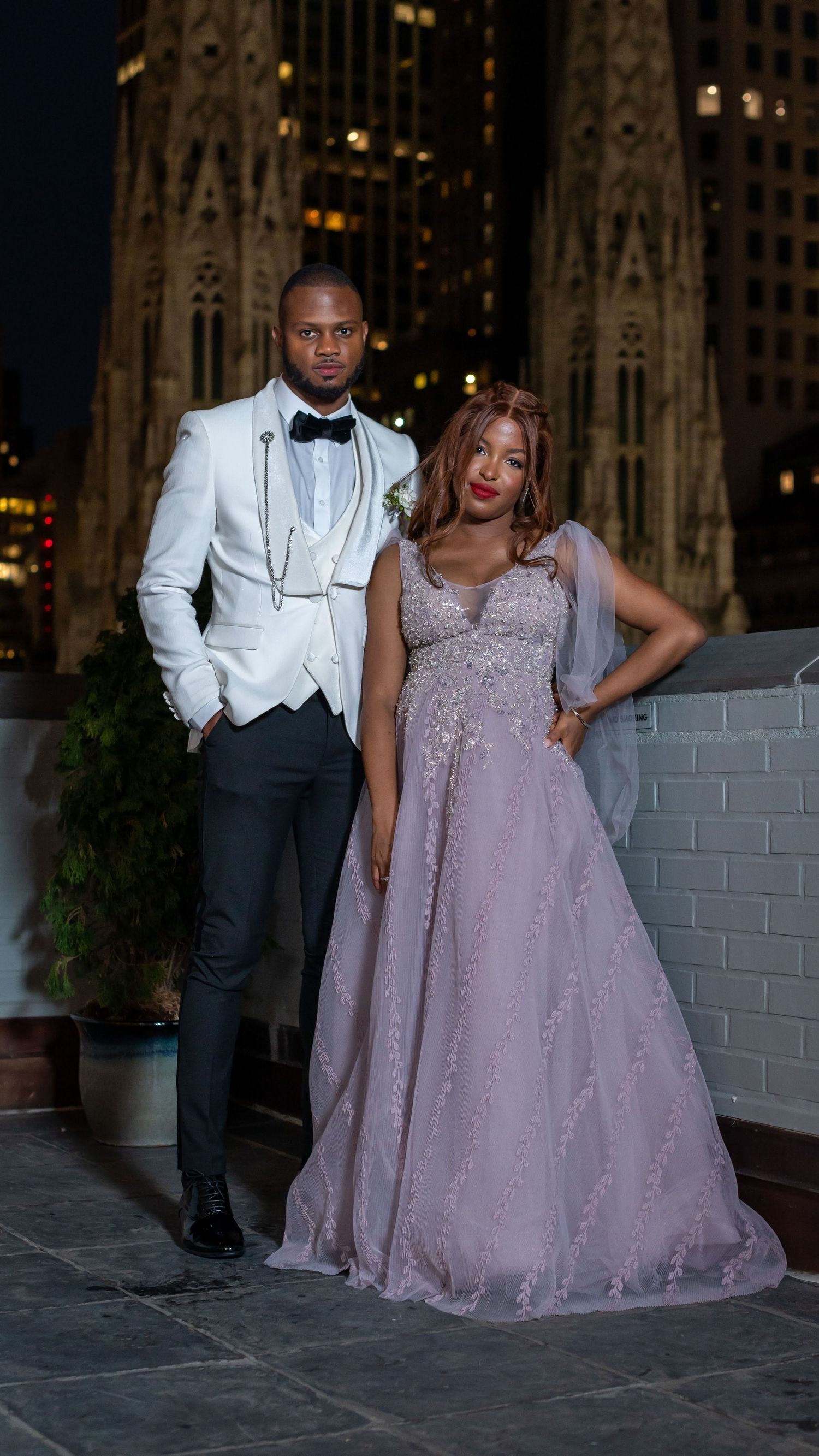 Formal couple in evening wear posing against gothic architecture at night.